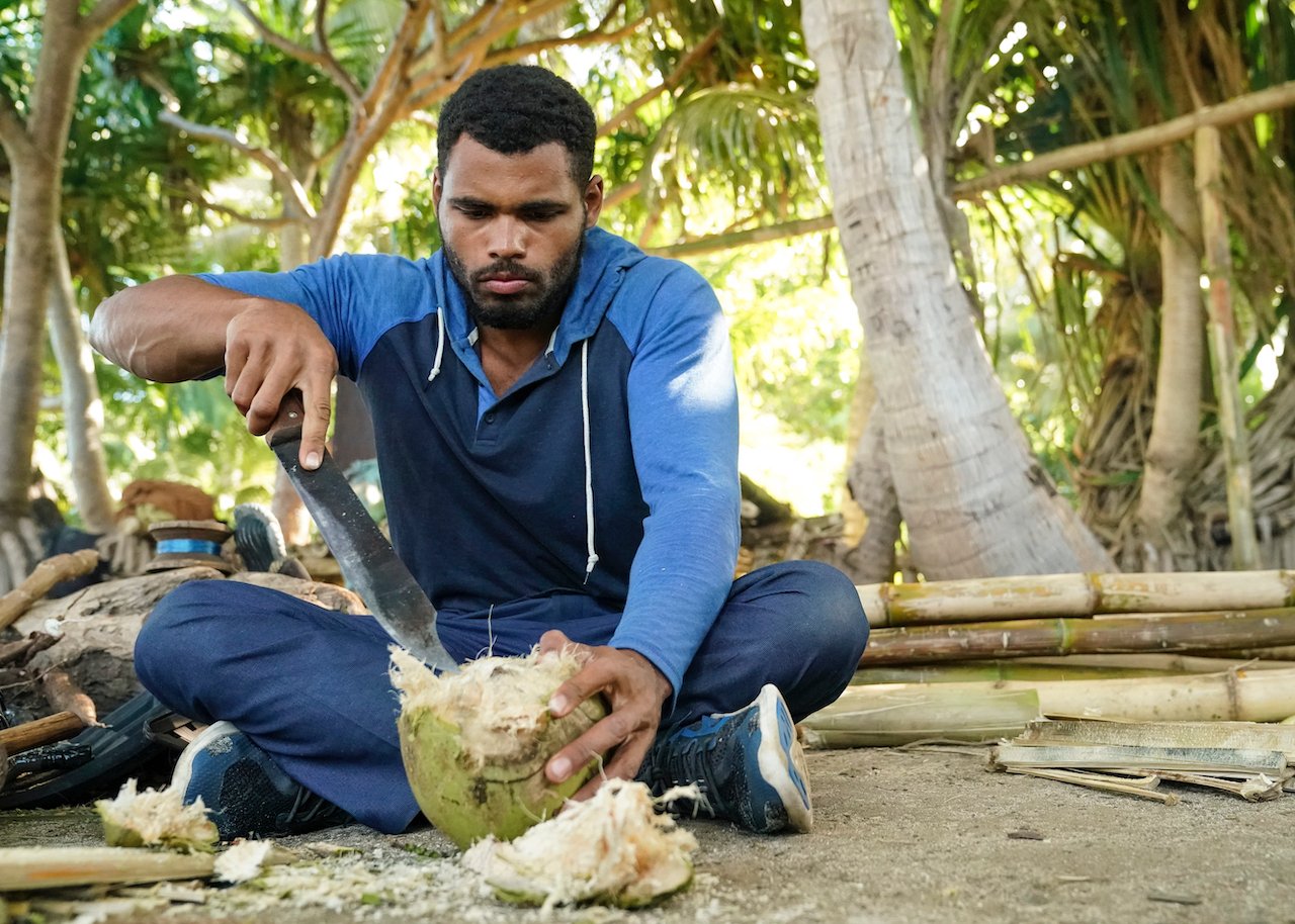 Ryan Medrano cuts a coconut with a machete while sitting on the ground on 'Survivor 43'.