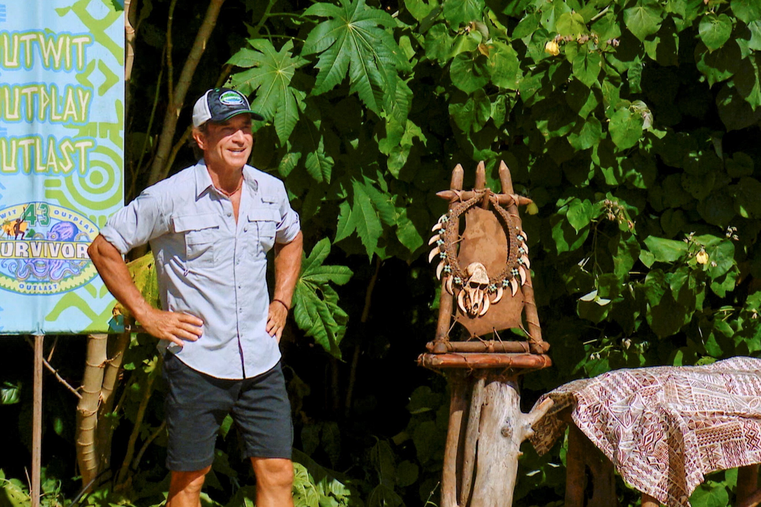 Jeff Probst, who will host the 'Survivor 44' premiere, wears a light blue button-down shirt, dark gray shorts, and a black, white, and green 'Survivor' hat. Probst stands next to the immunity necklace on the beach during 'Survivor 43.'