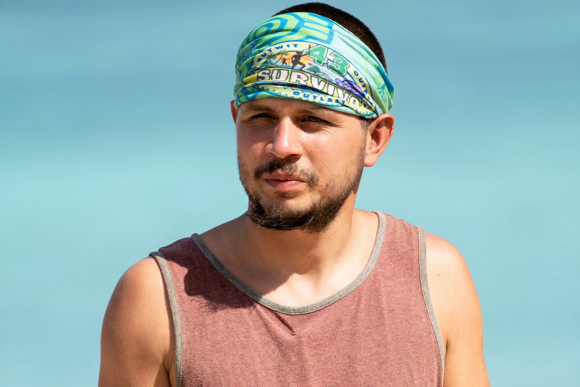 Jesse Lopez, who stars in 'Survivor' Season 43 on CBS, wears a faded red tank top and his blue 'Survivor' buff on his head.