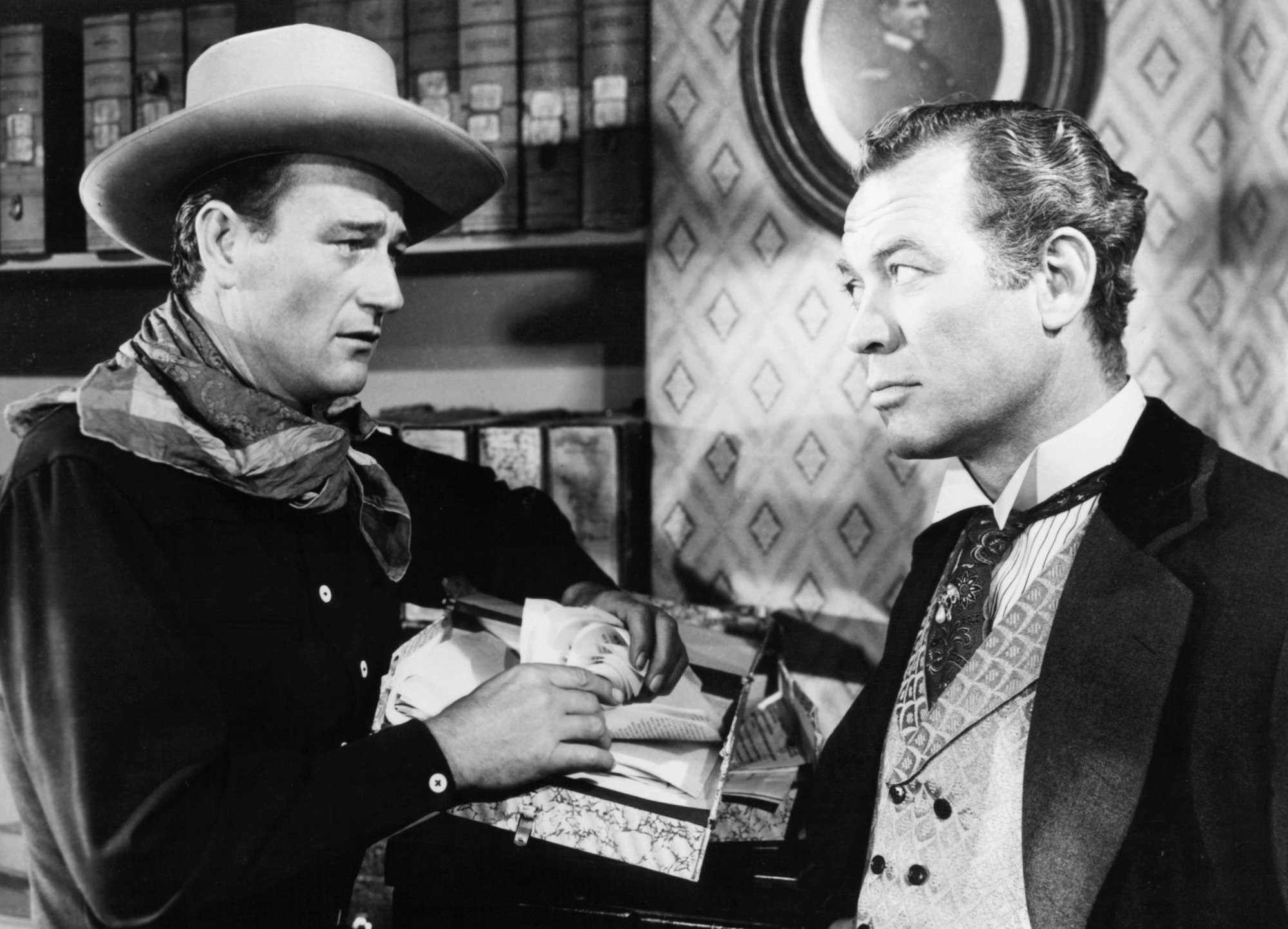 'Tall in the Saddle' John Wayne and Ward Bond. They're looking at each other in the eyes wearing Western costumes in front of a bookcase