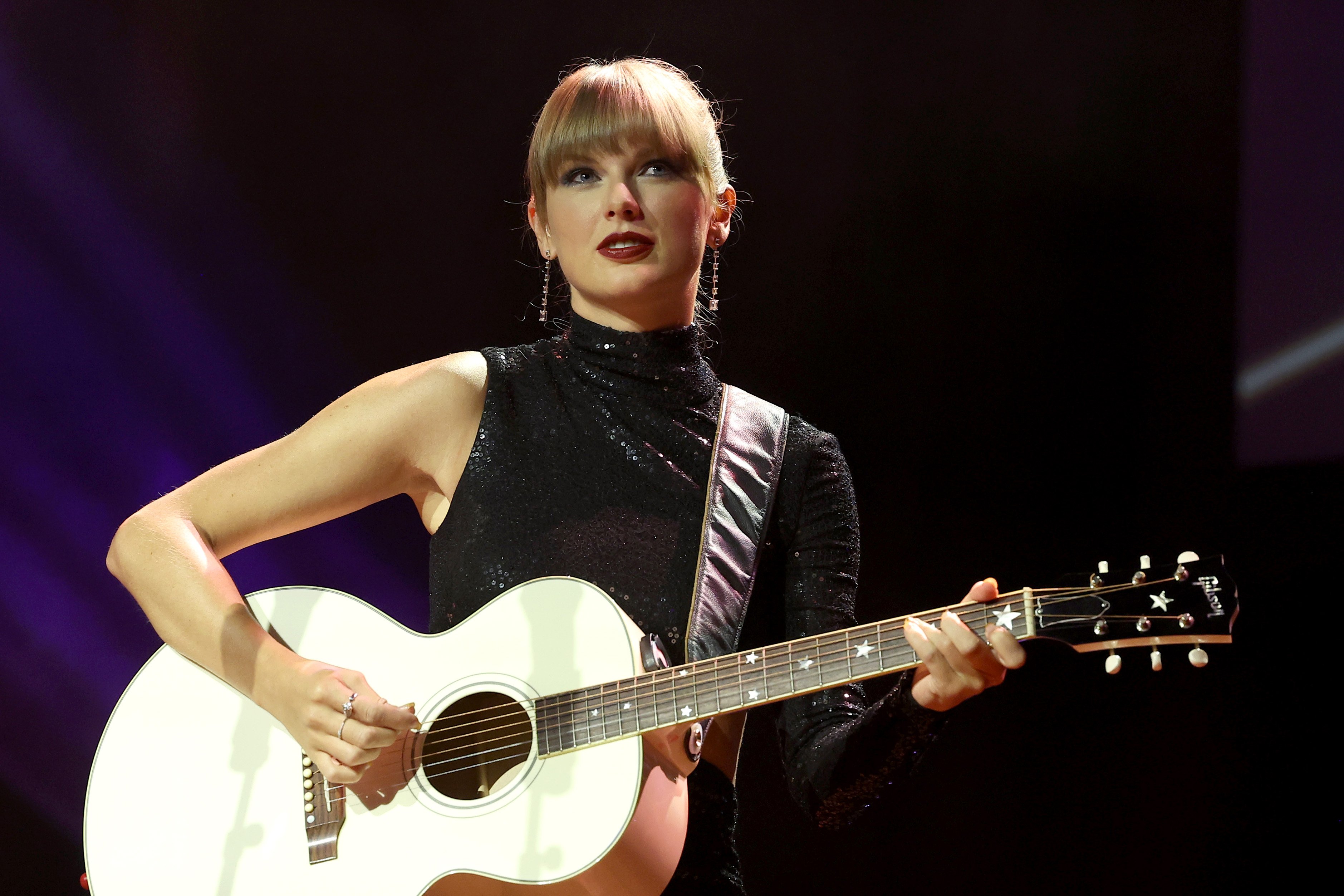Taylor Swift wears a black shirt while playing a white acoustic guitar
