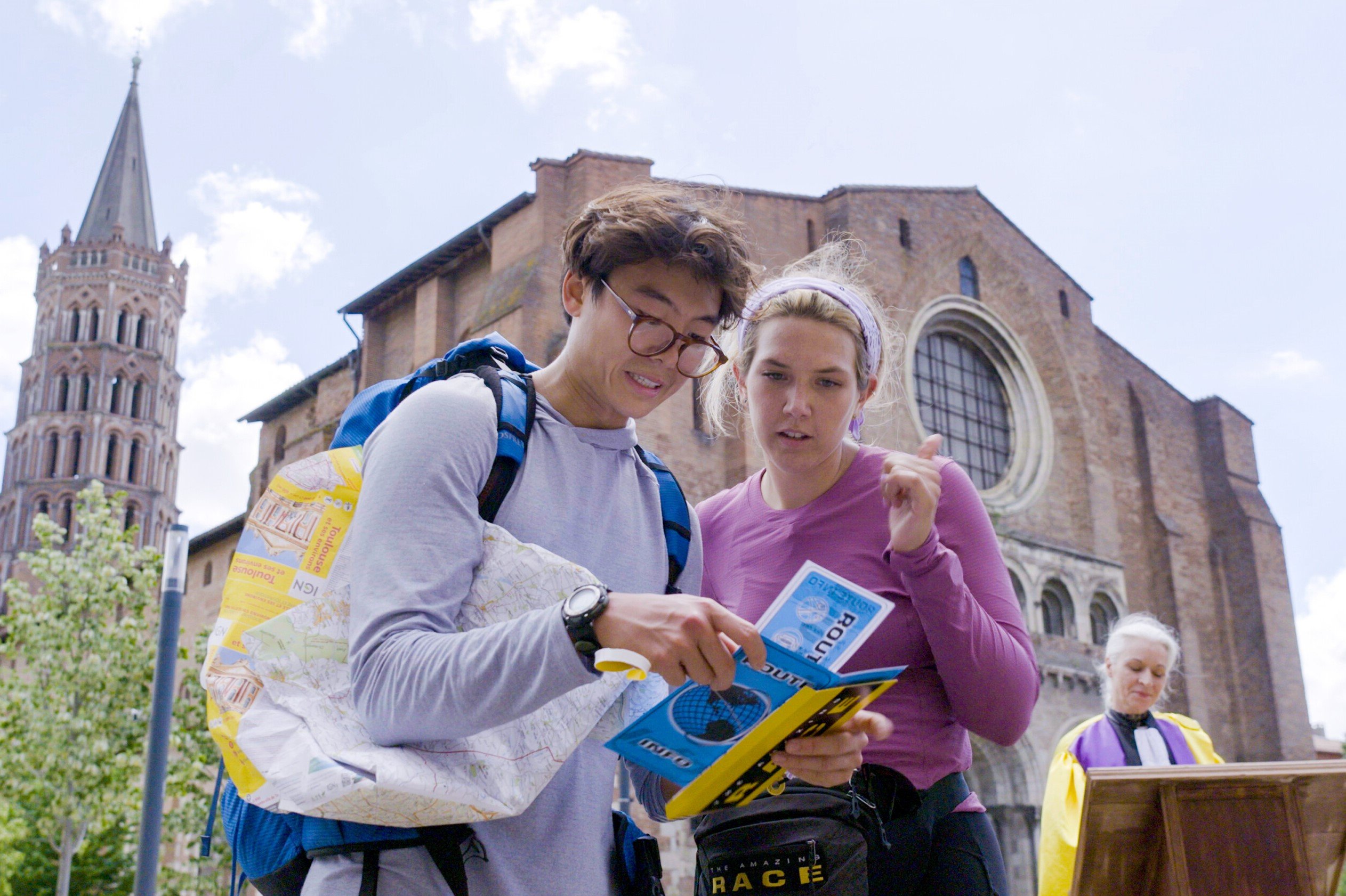 Derek Xiao and Claire Rehfuss, who will continue racing in 'The Amazing Race 34' Episode 9, read a clue. Derek wears a light blue hoodie. Claire wears a purple long-sleeved shirt and black leggings.