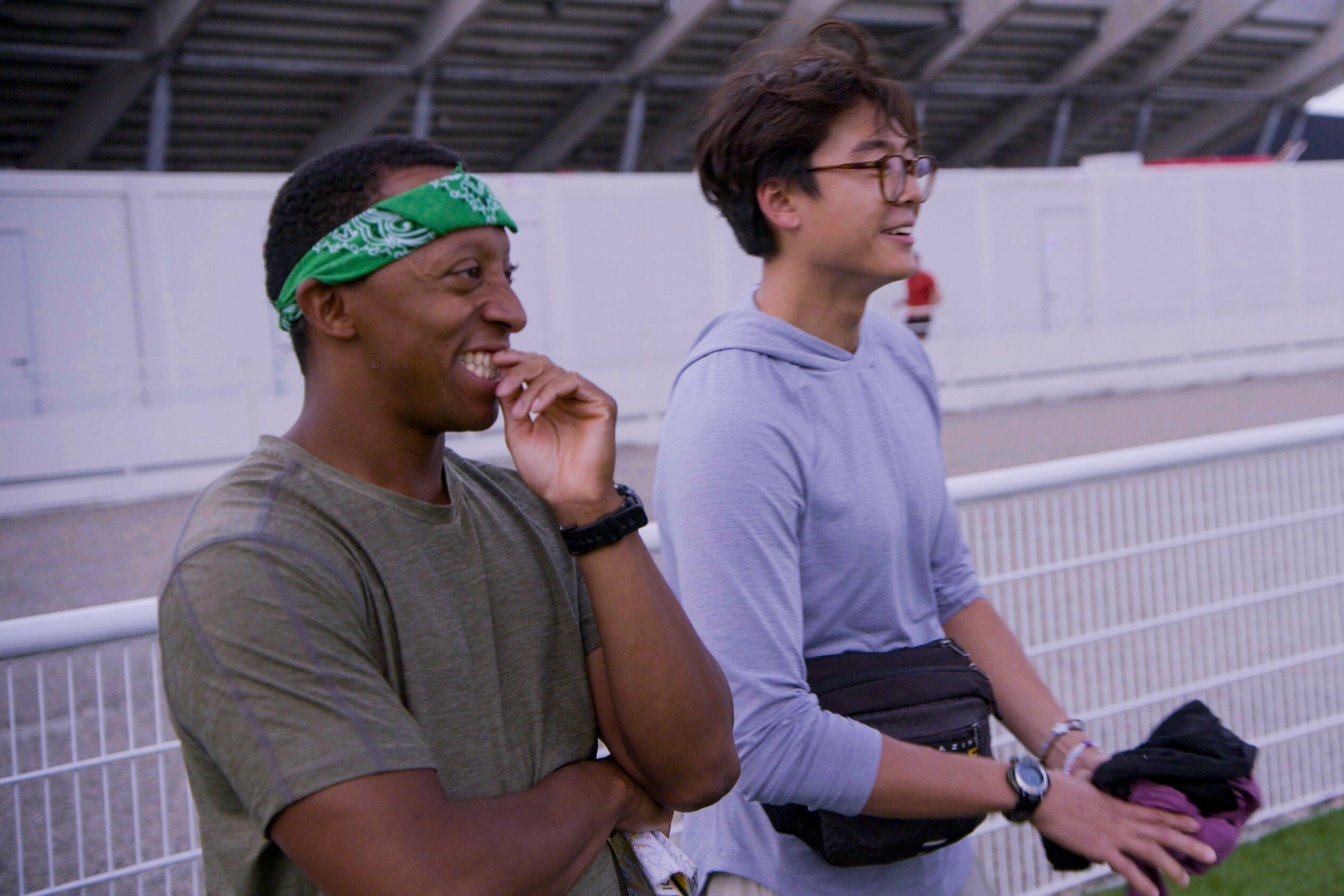 Michael Craig and Derek Xiao, who star in 'The Amazing Race' Seasons 34 on CBS, watch as their partners complete a Roadblock. Michael wears an olive green shirt and green bandana. Derek wears a light blue hoodie and black fanny pack.