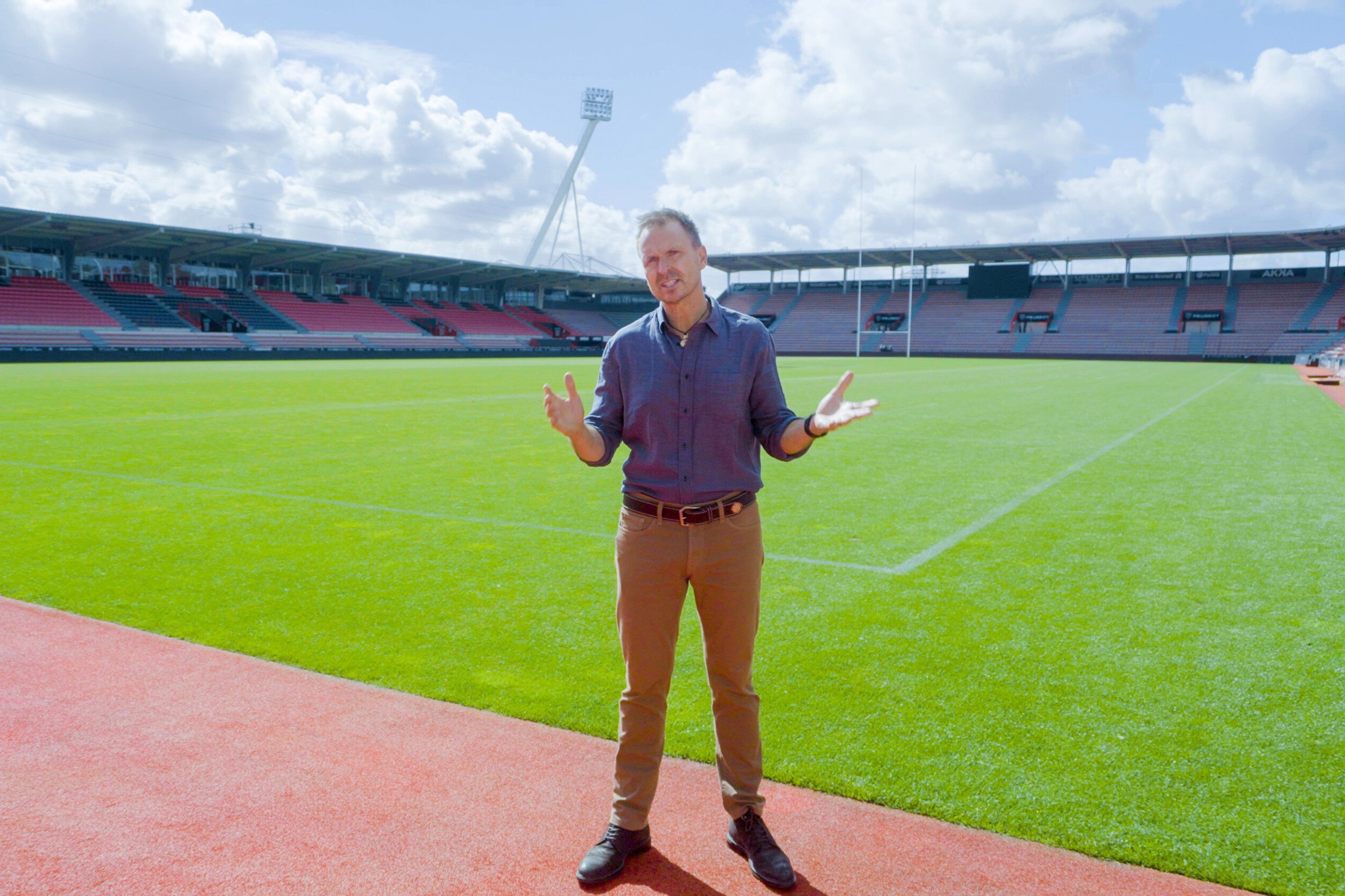Phil Keoghan, who people can watch host 'The Amazing Race' Season 34 on CBS, stands on a sports field wearing a blue button-down shirt and tan pants.