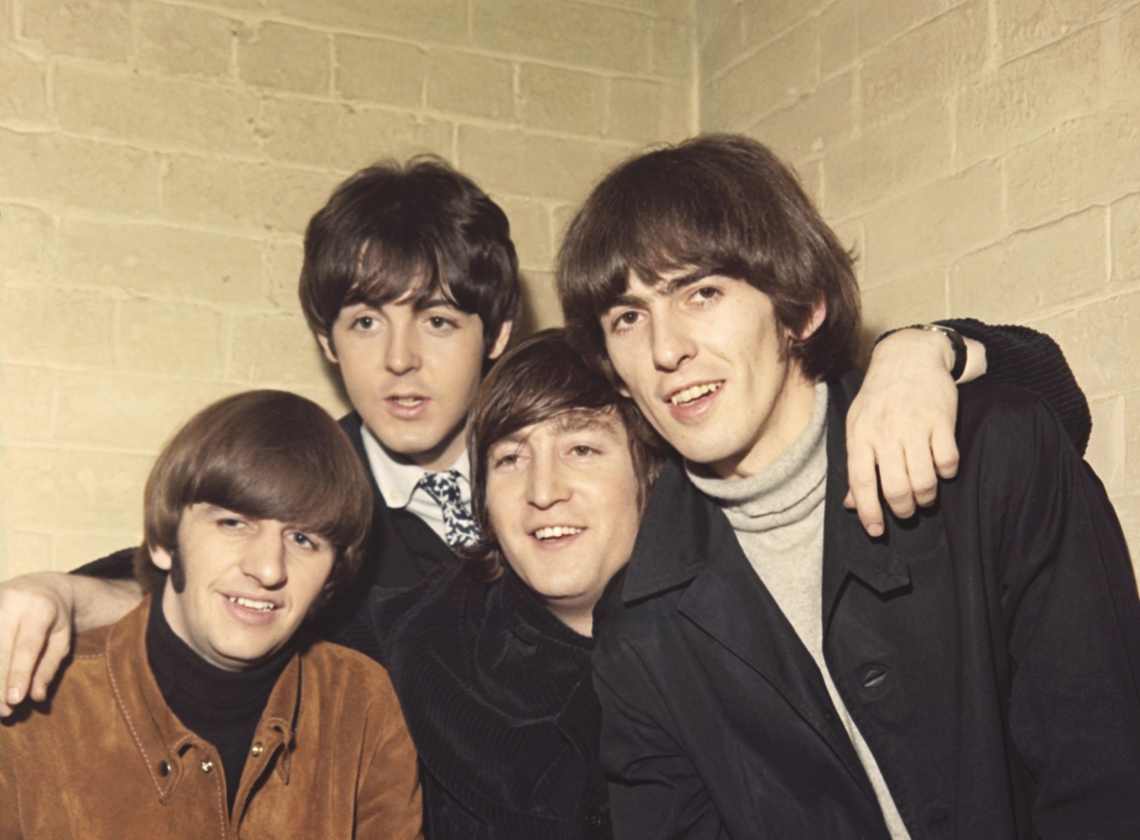 The Beatles, Ringo Starr, Paul McCartney, John Lennon, and George Harrison, pose together in front of a white brick wall.