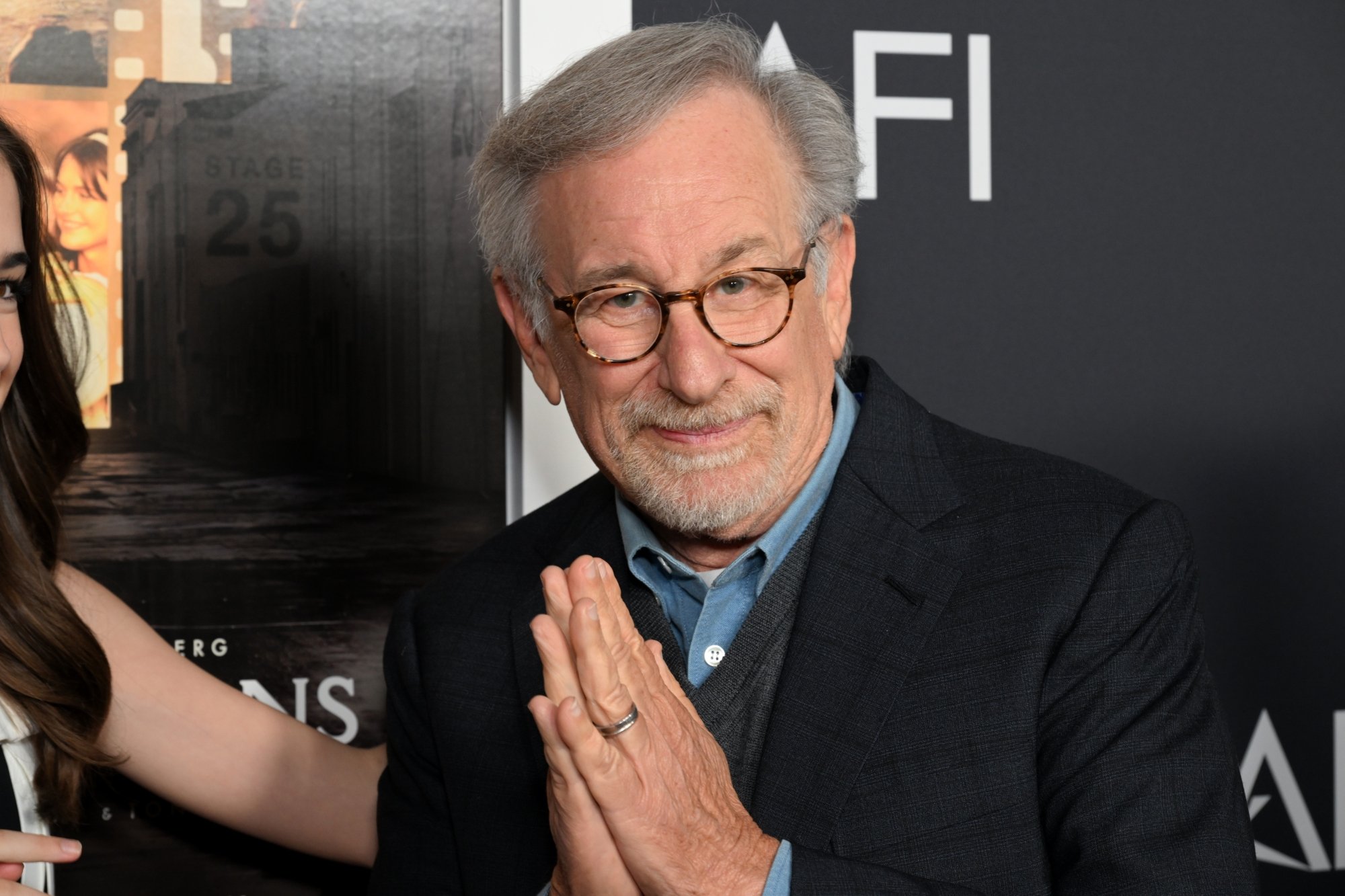'The Fabelmans' filmmaker Steven Spielberg. He has his hands clasped together, wearing a suit jacket in front of the AFI FEST step-and-repeat