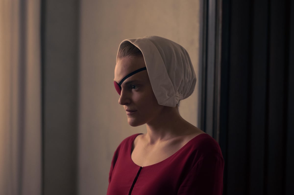 Madeline Brewer as Janine in The Handmaid's Tale. Janine wears a red dress and eye patch and a white bonnet.