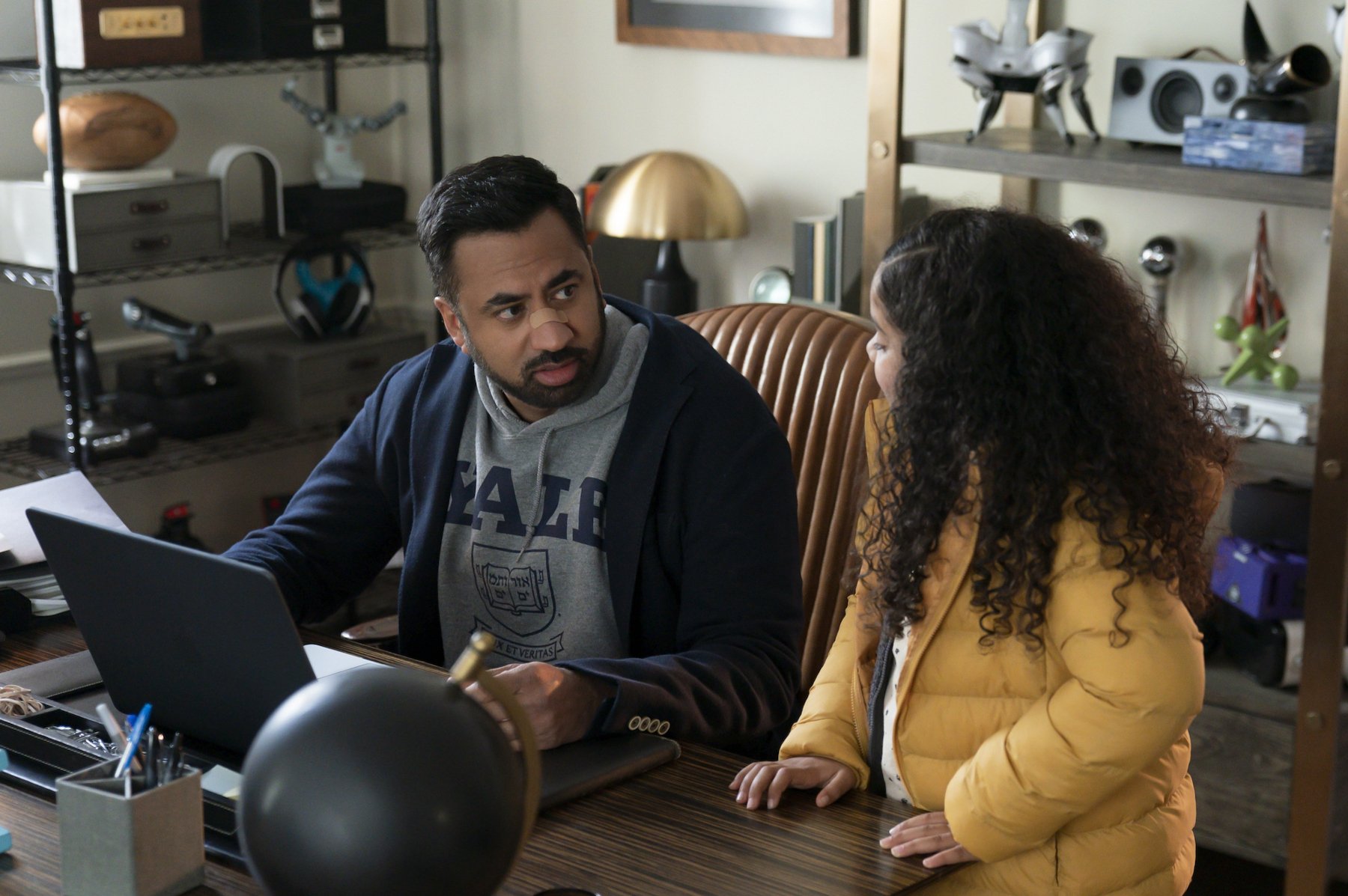 Kal Penn and Rupali Redd as Simon and Grace in 'The Santa Clauses' for our article previewing episode 3. They're sitting at a desk and looking at one another. Simon has a bandaid on his nose.