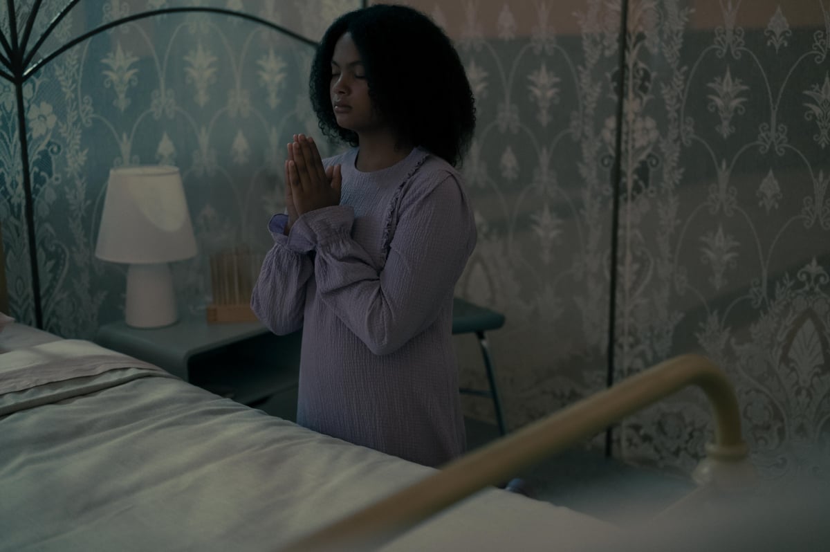 The Testaments fans should expect changes from the book. Hannah kneels by her bed and prays, wearing a purple nightgown.