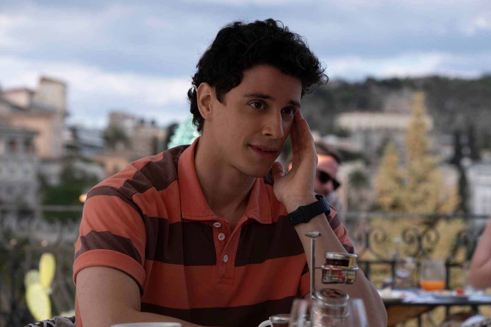 Adam DiMarco as Albie in 'The White Lotus' Season 2. He's sitting at a table and wearing a pink and blue shirt.