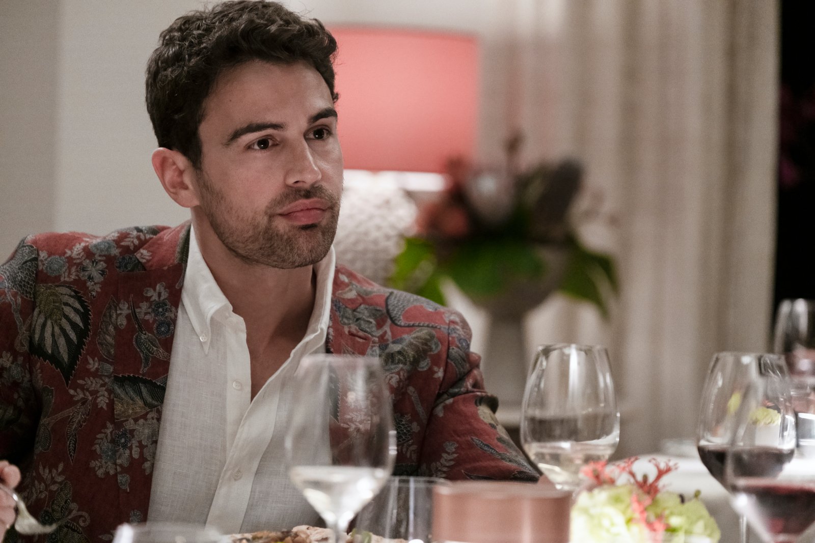 Theo James as Cameron in 'The White Lotus' Season 2. He's wearing a white shirt, red and green blazer, and sitting at a table.