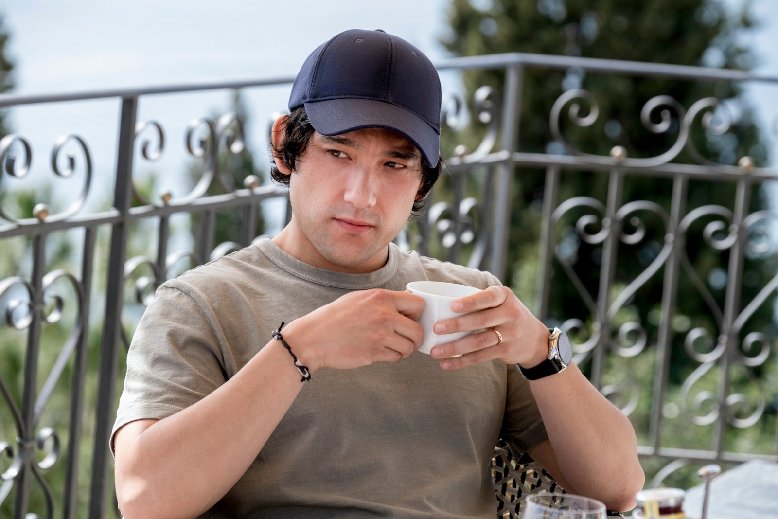 Will Sharpe as Ethan in 'The White Lotus' Season 2. He's sitting down, wearing a dark blue baseball cap, and holding a white mug.