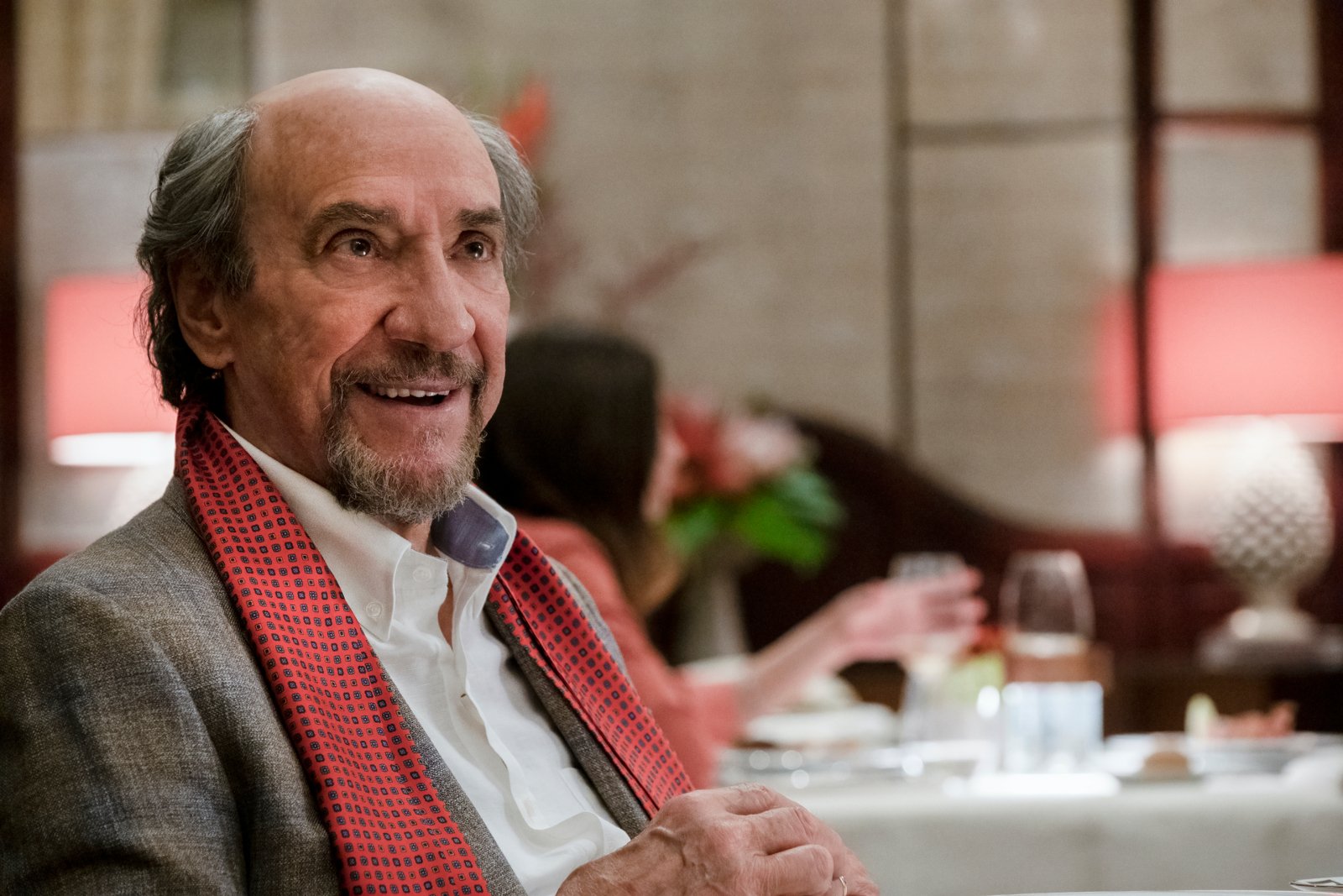 F. Murray Abraham as Bert Di Grasso in 'The White Lotus' Season 2 for our article about his Hades and Persephone comment. He's sitting in a restaurant and wearing a white shirt, grey suit, and pink tie around his shoulders.