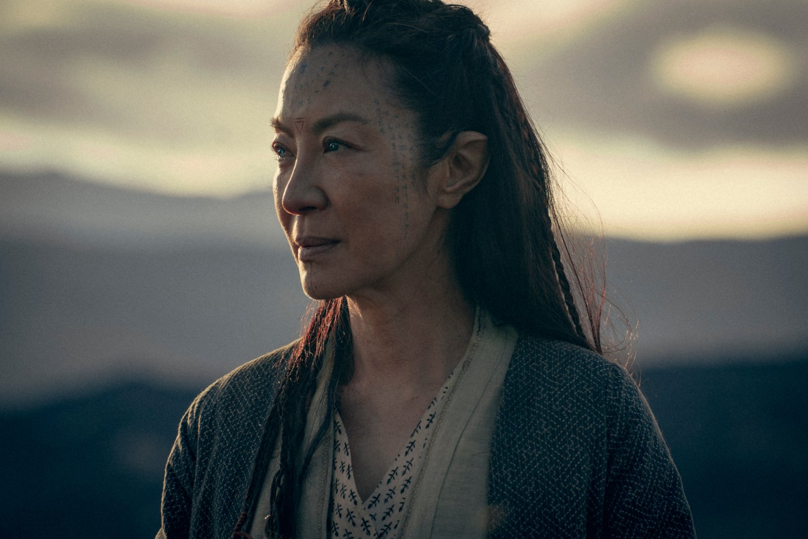 Michelle Yeoh in 'The Witcher: Blood Origin' for our article about its release date, trailer, cast, and more. She's looking at something off-screen, and the sky is cloudy behind her.
