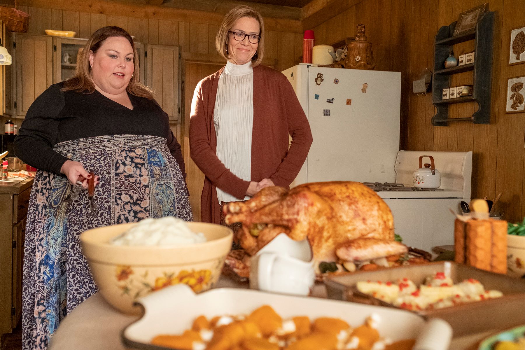 Chrissy Metz and Mandy Moore, in character as Kate and Rebecca in 'This Is Us' Season 6 Episode 7, admire their Thanksgiving dinner. Kate wears a black long-sleeved top and long blue paisley skirt. Rebecca wears a red-brown cardigan over a white turtleneck.