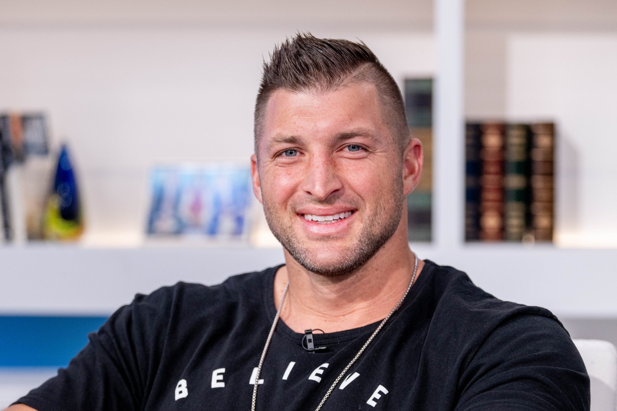 Tim Tebow, who 'Saturday Night Live' made a sketch about. He's smiling and wearing a black t-shirt and a necklace in front of a bookshelf.