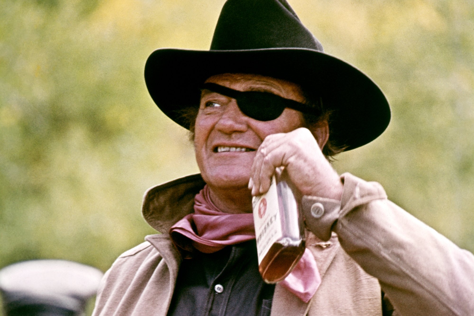 'True Grit' John Wayne as Rooster Cogburn smiling, holding up a bottle of booze. He's wearing a cowboy hat and wearing an eyepatch over his eye.