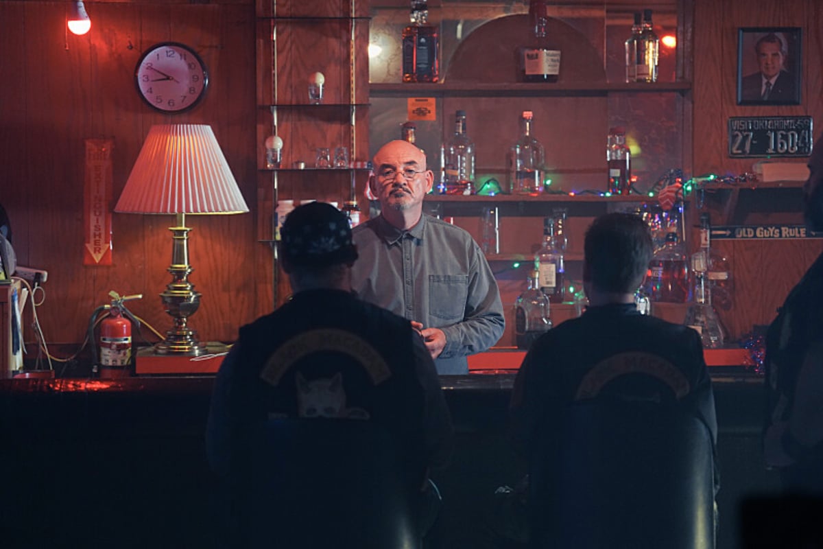 Ritchie Coster as Caolan Waltrip in Tulsa King. Caolan stands behind a bar with bikers seated in front of him.