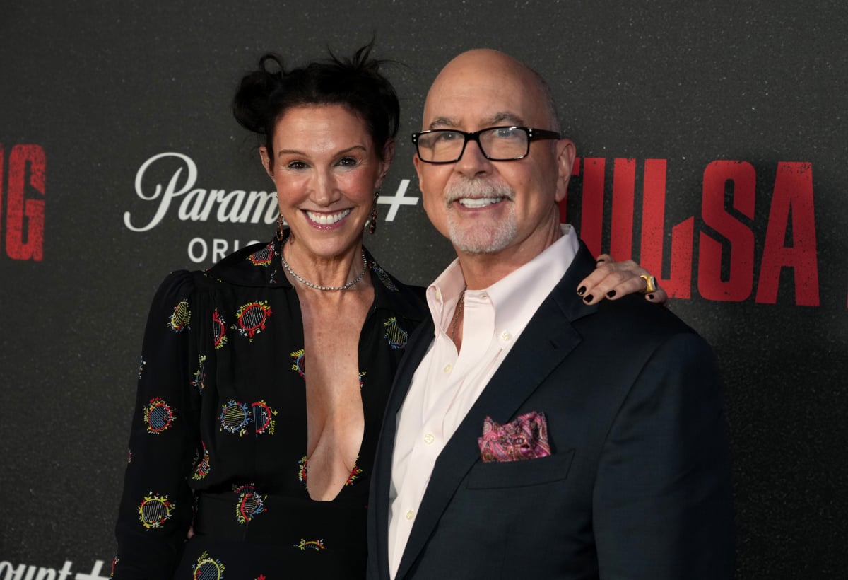 Terence Winter attends the Tulsa King premiere with his wife, Rachel. He is wearing a black suit jacket and white button-down shirt and she is wearing a patterned blazer. 