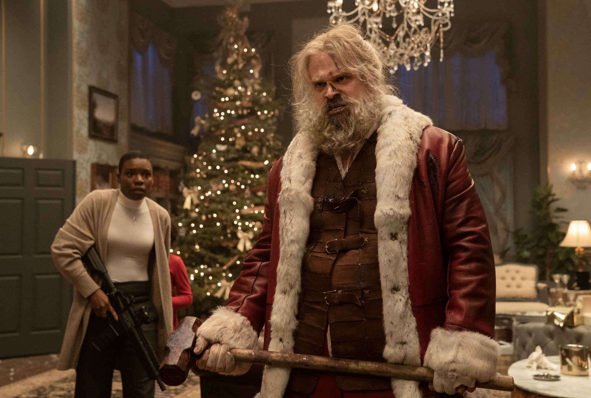 'Violent Night' Alexis Louder as Linda and David Harbour as Santa Claus. Linda is holding a gun and Santa is holding a sledgehammer. He's all beat up standing in front of a Christmas tree.