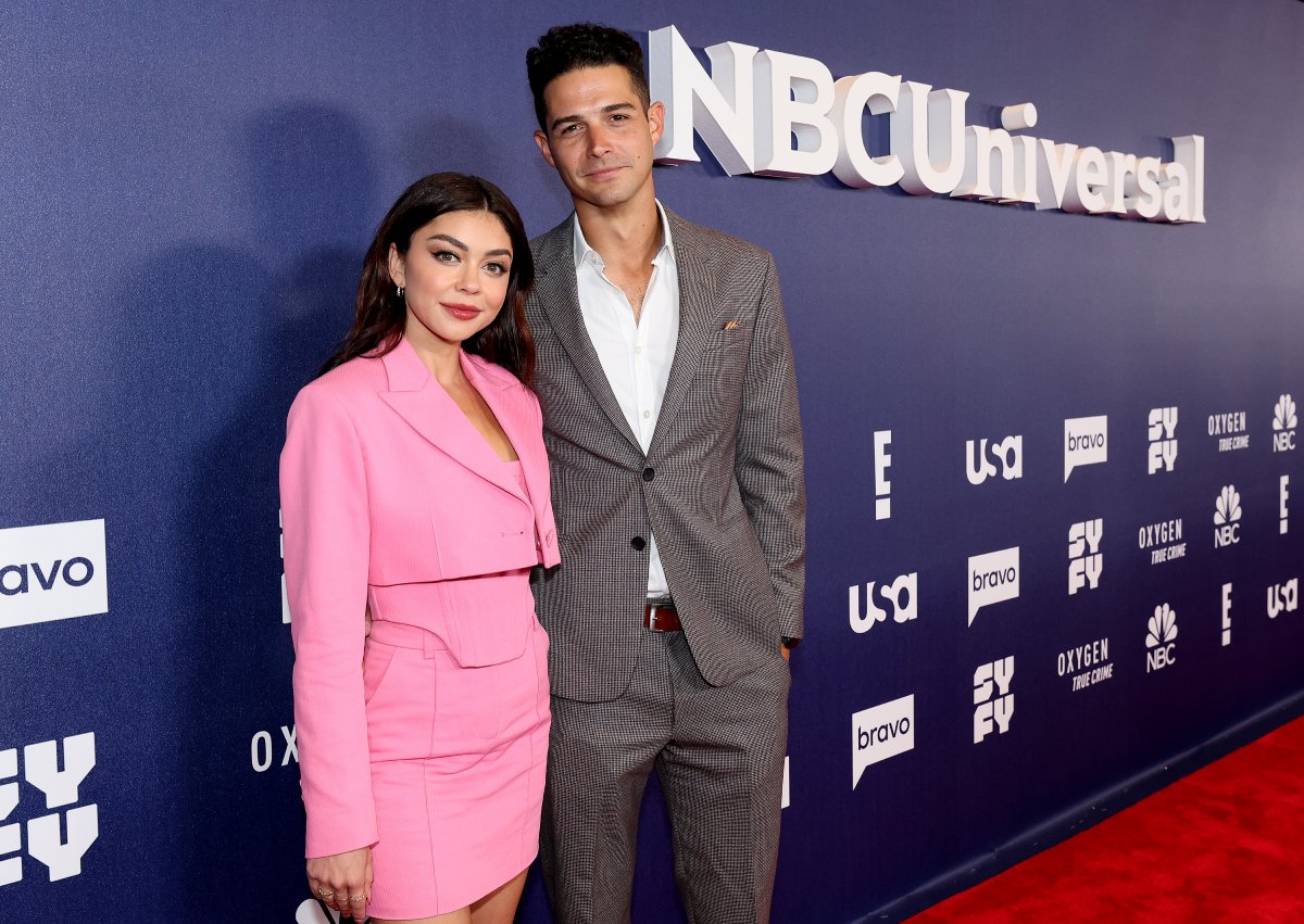 Wells Adams and Sarah Hyland pose for a photo. Hyland wears an all-pink outfit and Adams wears a gray suit and white button-down shirt. 