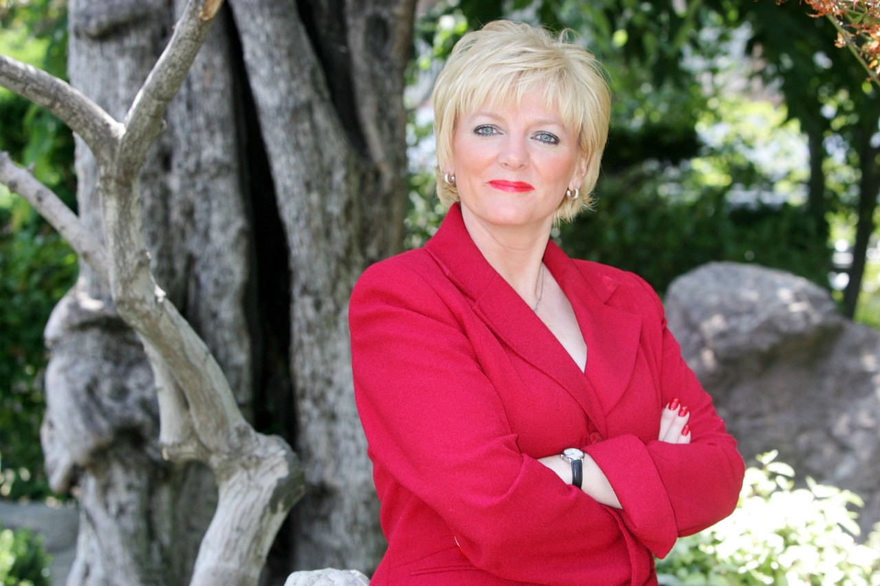 Alison Arngrim, Nellie Oleson from Little House on the Prairie, stands near a tree in a red blazer with her arms folded.