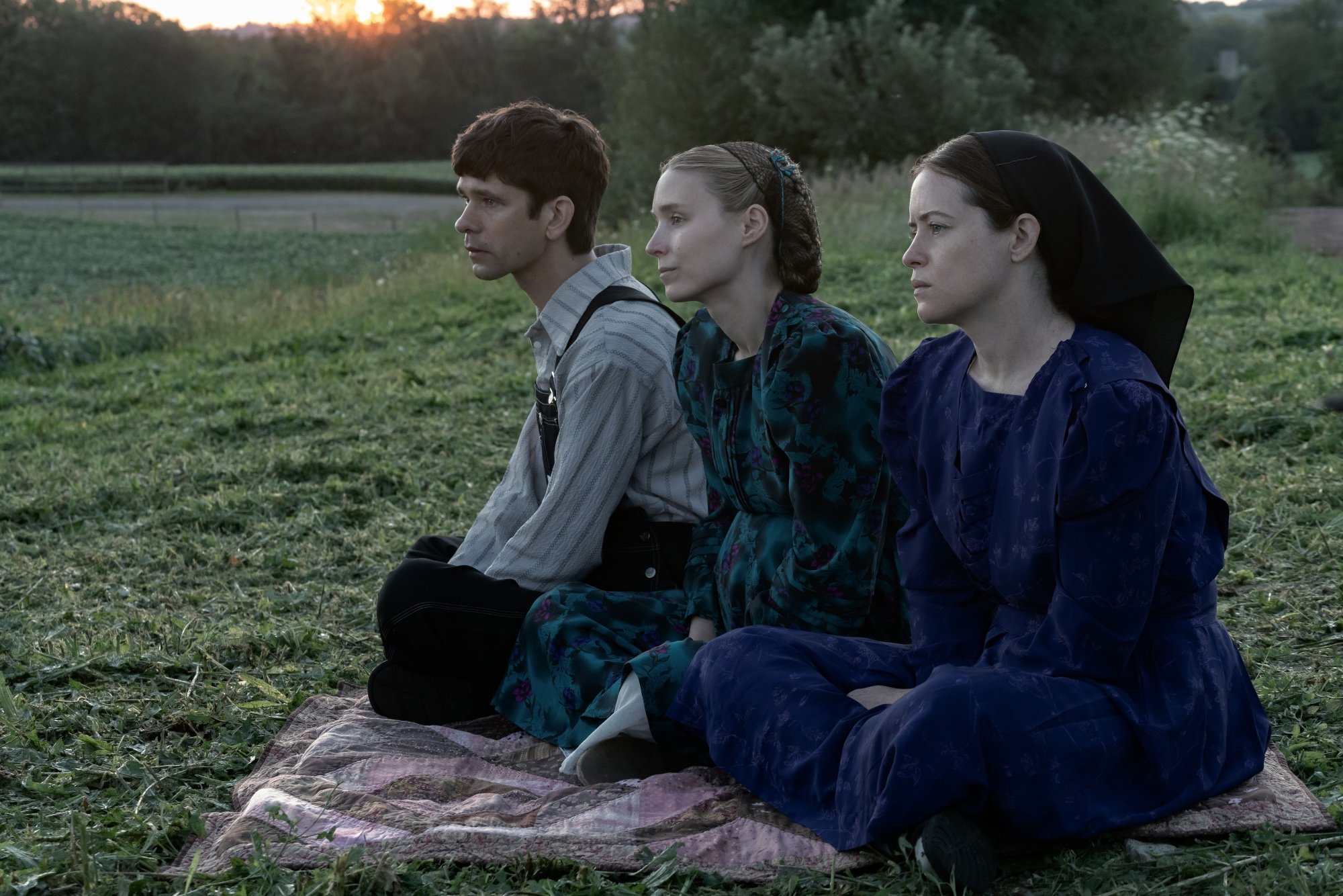 'Women Talking' Ben Whishaw stars as August, Rooney Mara as Ona and Claire Foy as Salome sitting on a blanket in a field of grass looking ahead with a serious look on their faces