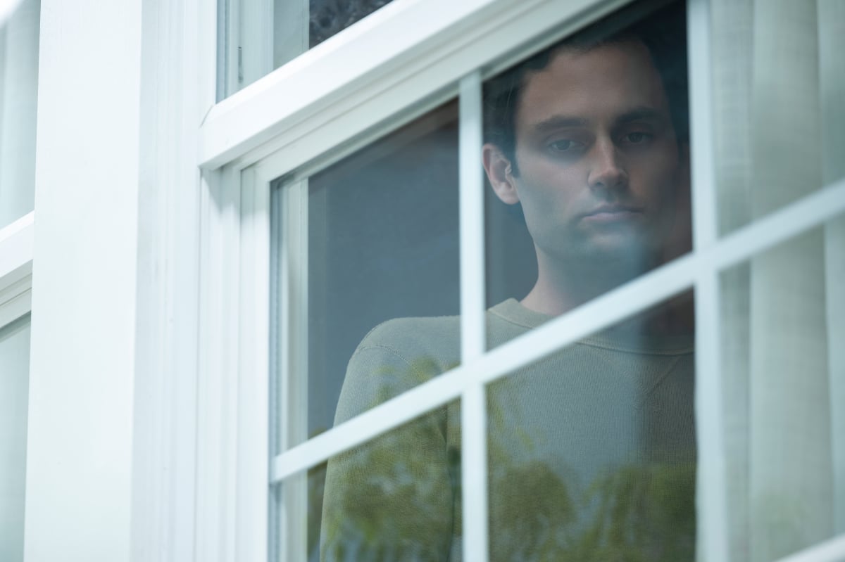 Joe Goldberg returns for You Season 4. Joe wears a green long-sleeved shirt and stares out of a window.