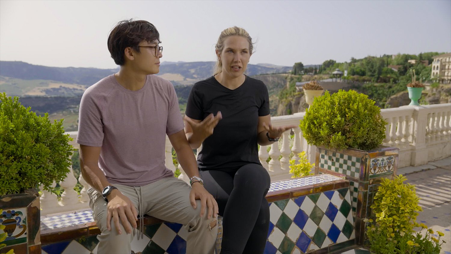 The Amazing Race 34: Derek Xiao sits on a ledge in a purple T-shirt as he looks at Claire Rehfuss beside him, who wears a black T-shirt and talks with her hands spread out.