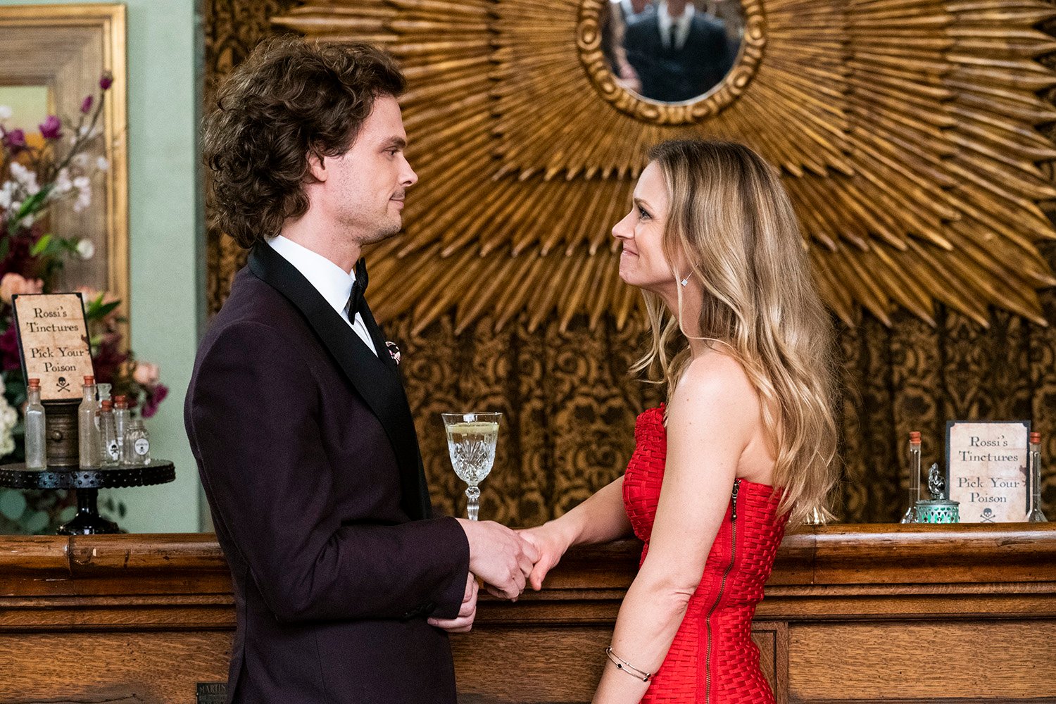 Criminal Minds stars Matthew Gray Gubler as Spencer Reid and A.J. Cook as JJ facing each other beside a bar and smiling while wearing a tuxedo and a red dress