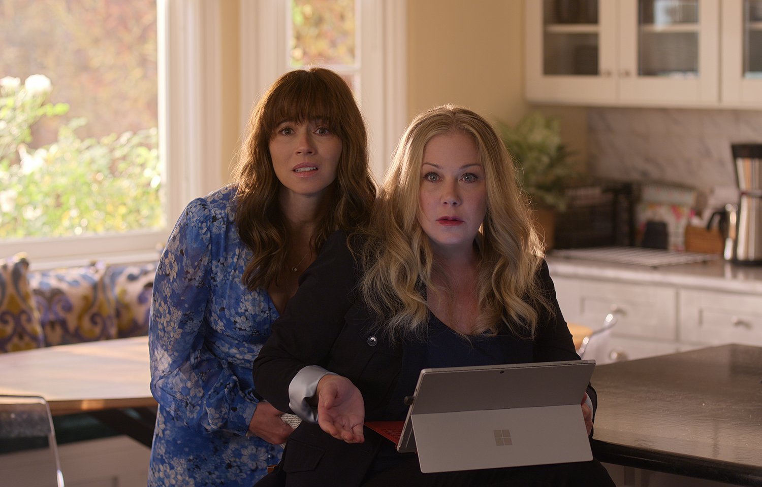 Dead to Me Season 3: Linda Cardellini's Judy Hale stands close to Christina Applegate's Jen Harding as she sits at a counter with a tablet.