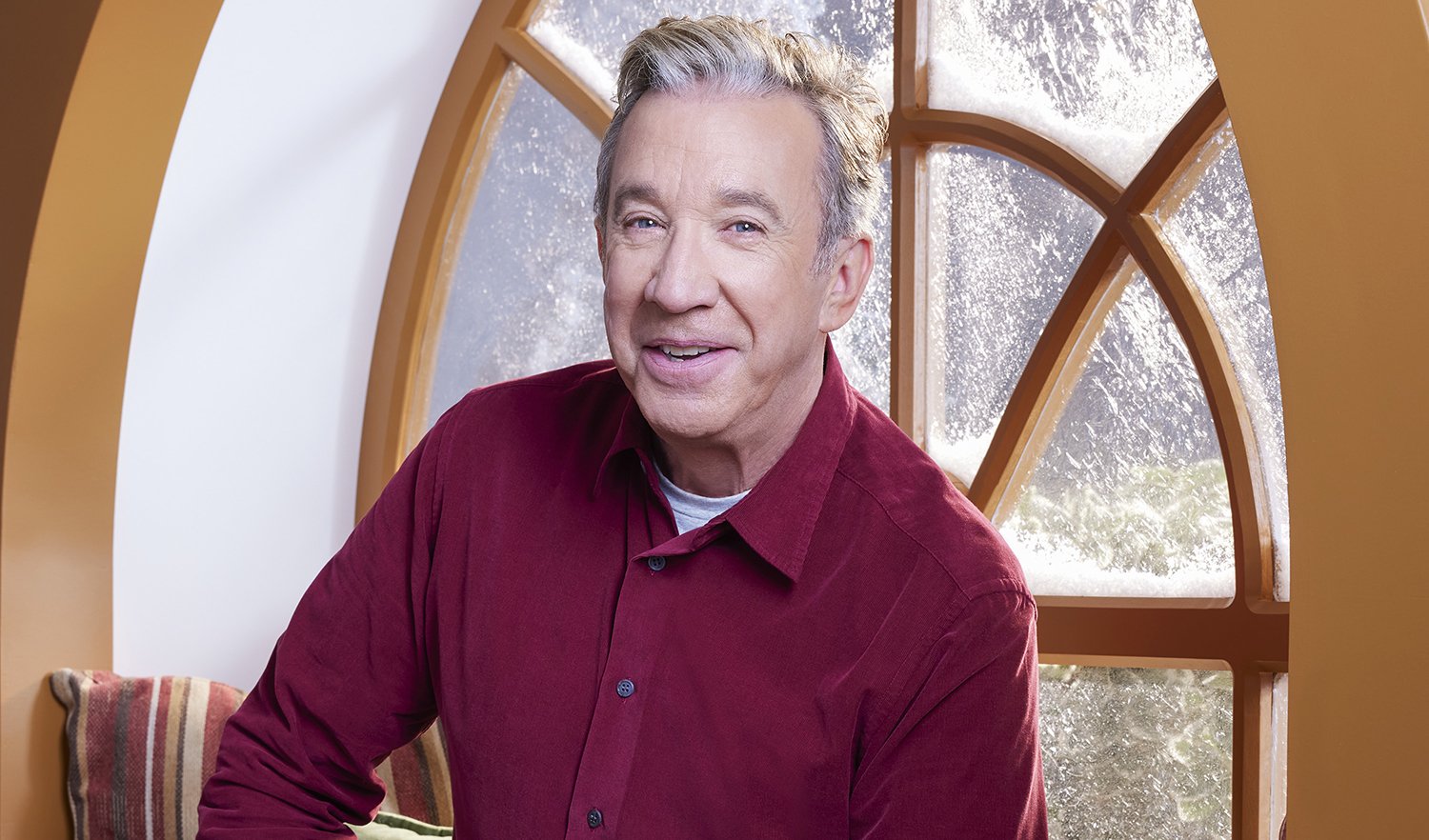The Santa Clause cast member Tim Allen sits by a frosty window and smiles while wearing a maroon button-down shirt.