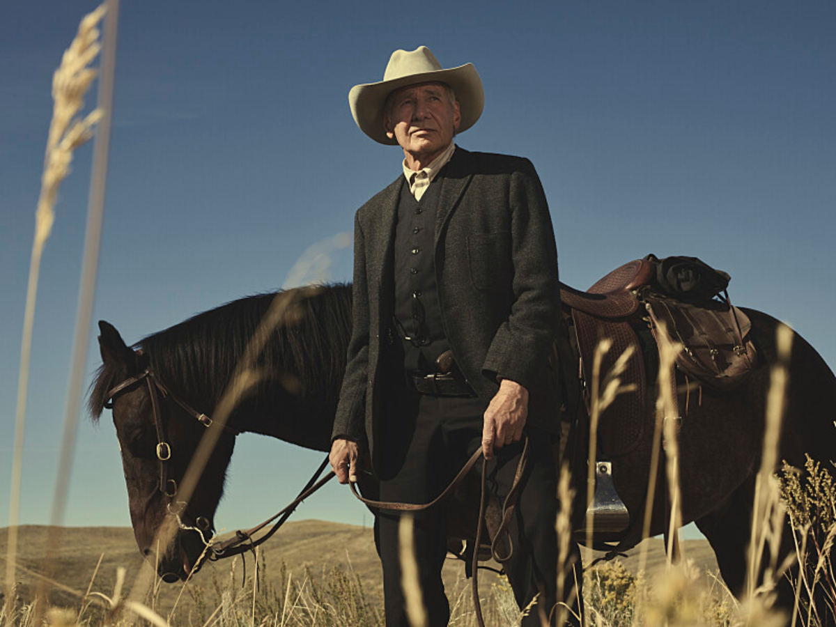 Harrison Ford as Jacob Dutton in Yellowstone. Jacob leads a horse. He is wearing an all-black outfit and a cream-colored cowboy hat.