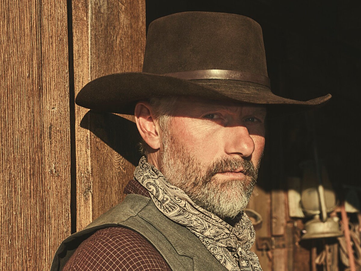 James Badge Dale plays John Dutton Sr. in '1923.' John wears a brown cowboy hat, scarf, vest, and shirt, and has a beard.