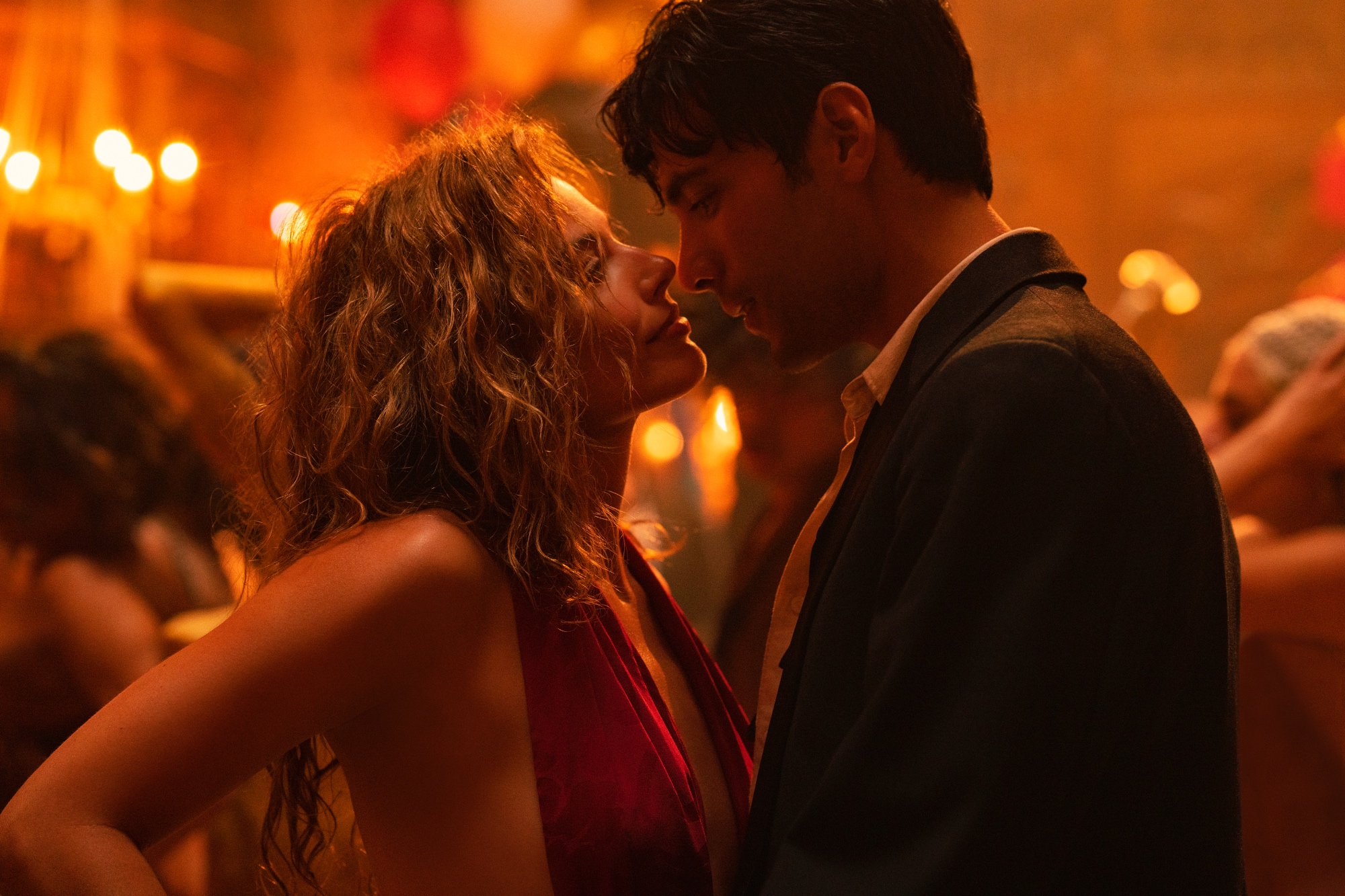 'Babylon' Margot Robbie as Nellie LaRoy and Diego Calva as Manny Torres looking into each other's eyes with their lips close. Robbie is wearing a red dress and Calva is wearing a suit.