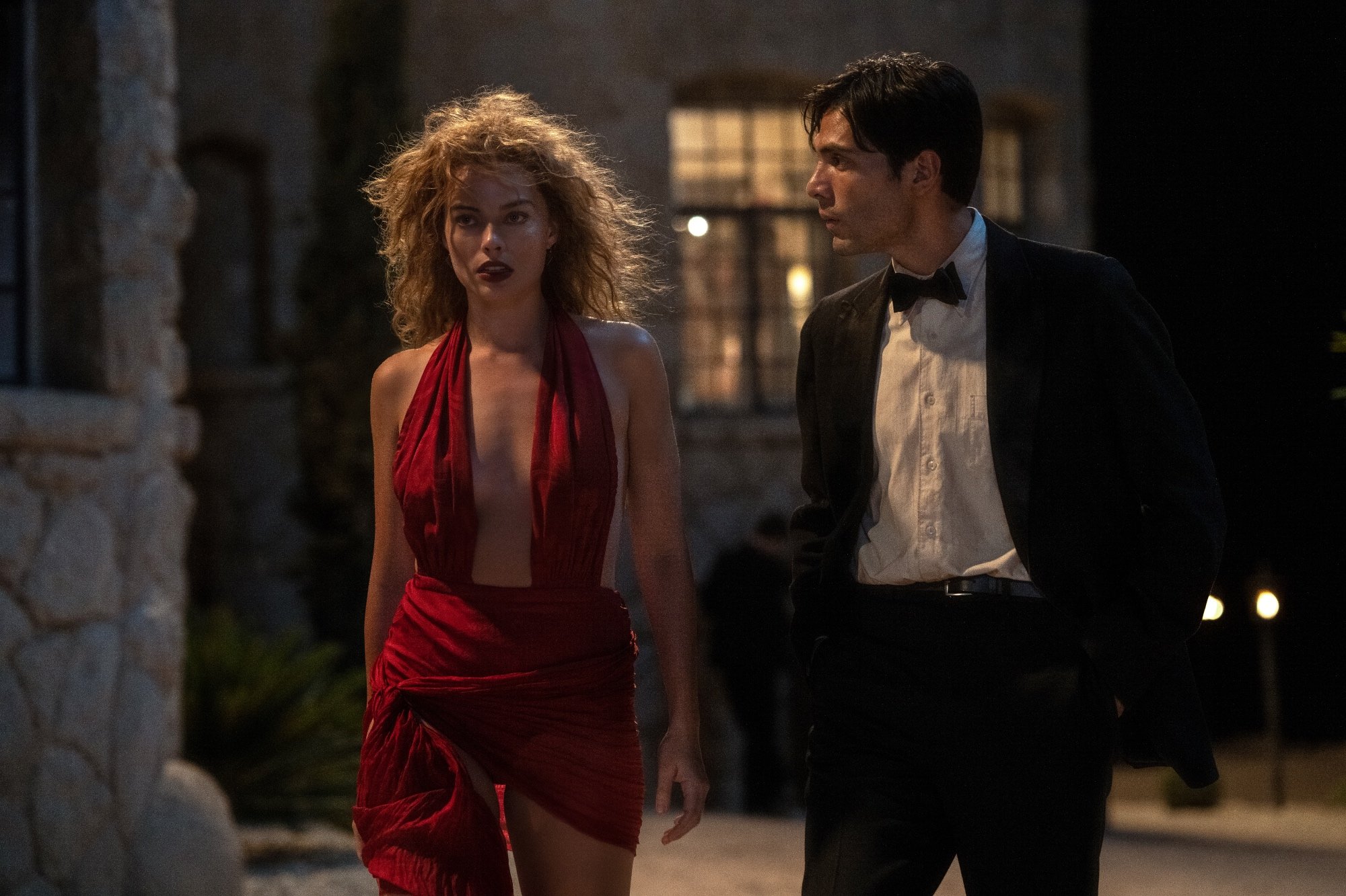 'Babylon' Margot Robbie as Nellie LaRoy and Diego Calva as Manny Torres walking outside of a house. She's wearing a red dress and he's wearing a black and white tux.