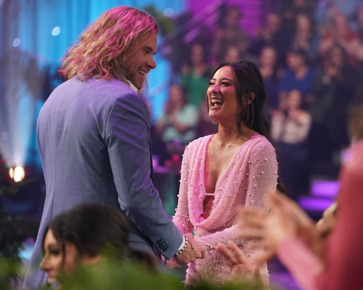 During the Bachelor in Paradise reunion, Jacob and Jill talk and laugh on stage. Jill is wearing a pink dress and Jacob is wearing a grey suit. 