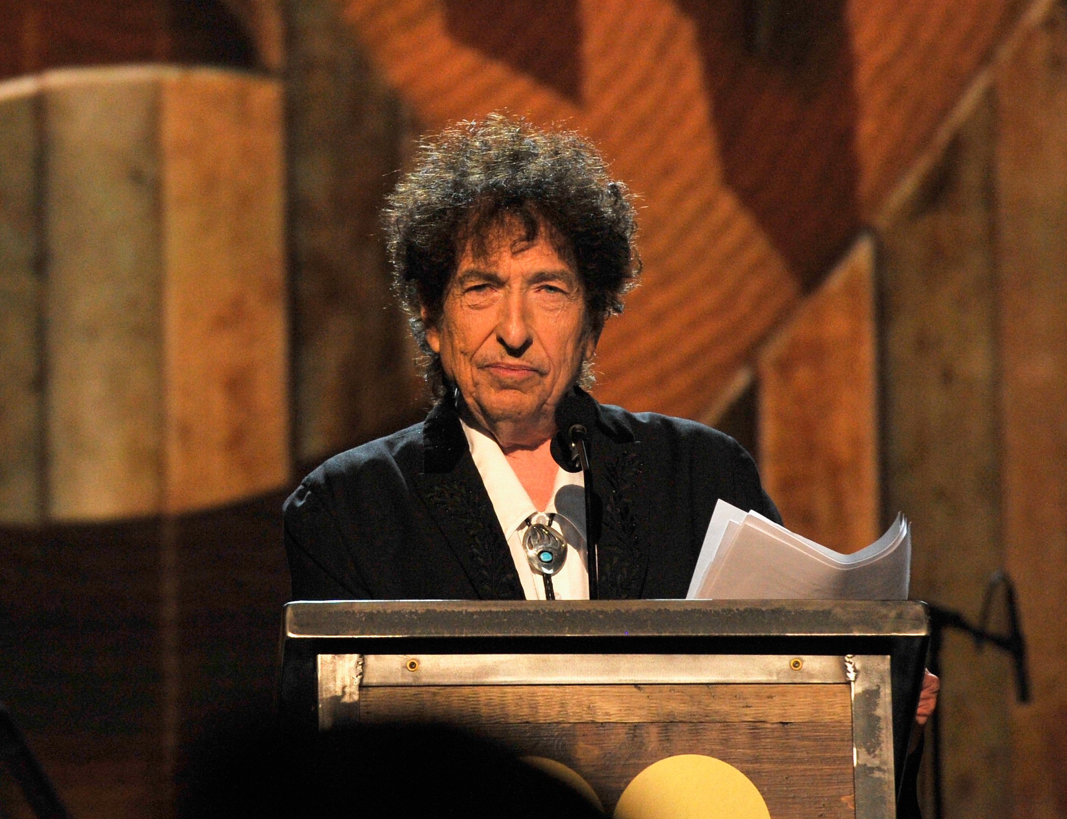 Bob Dylan stands at a podium and holds a stack of papers.