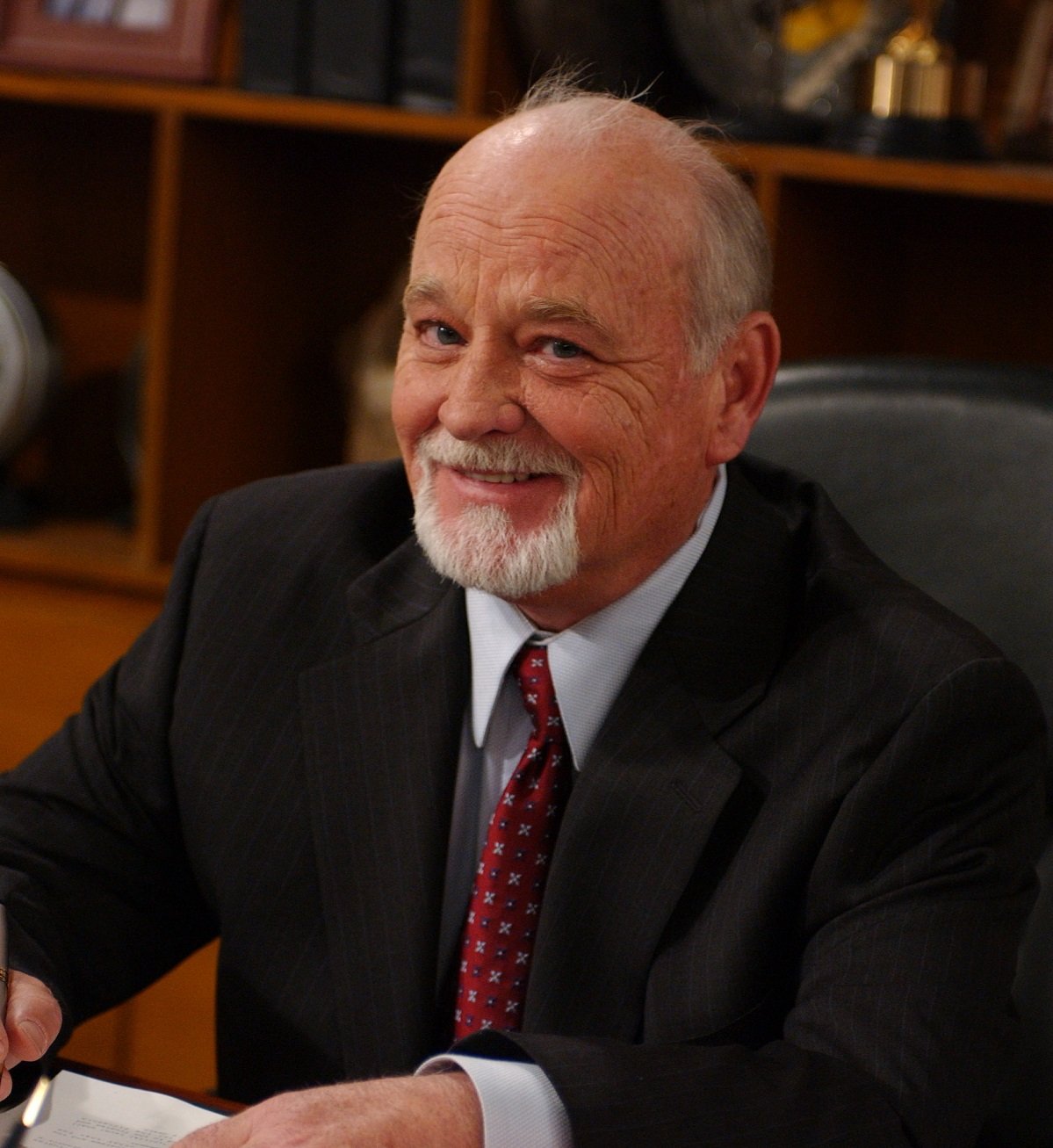 Actor Brain Doyle-Murray wearing a black suit, sitting at a desk on the set of the TV series 'Yes, Dear.'