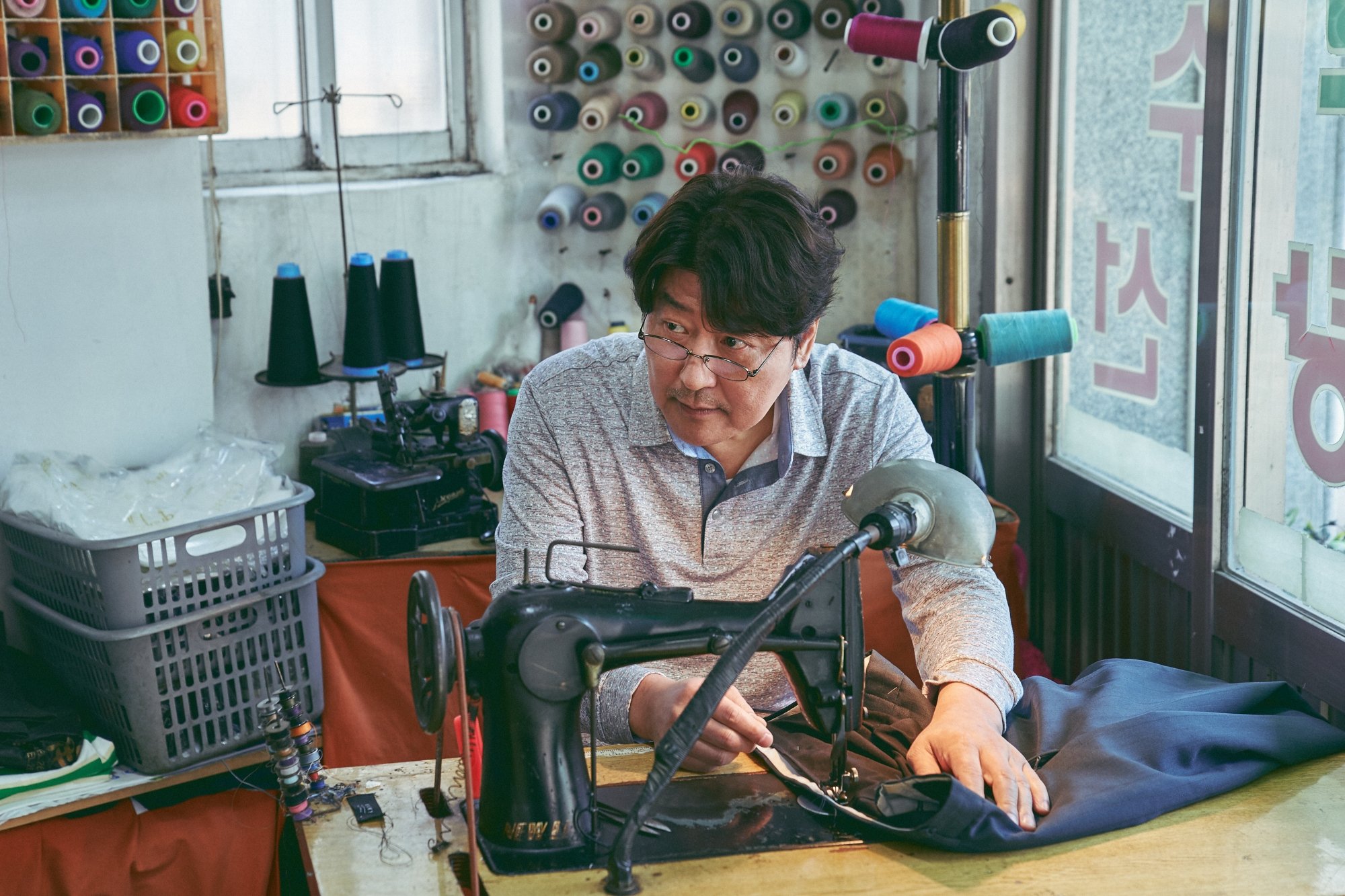 'Broker' Song Kang-ho as Sang-hyeon looking from under his glasses while he's working at a sewing machine