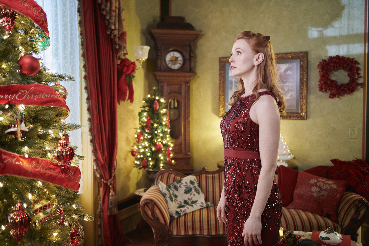 Alicia Witt wearing a red dress and looking at a Christmas tree in the Hallmark movie 'Christmas on Honeysuckle Lane'