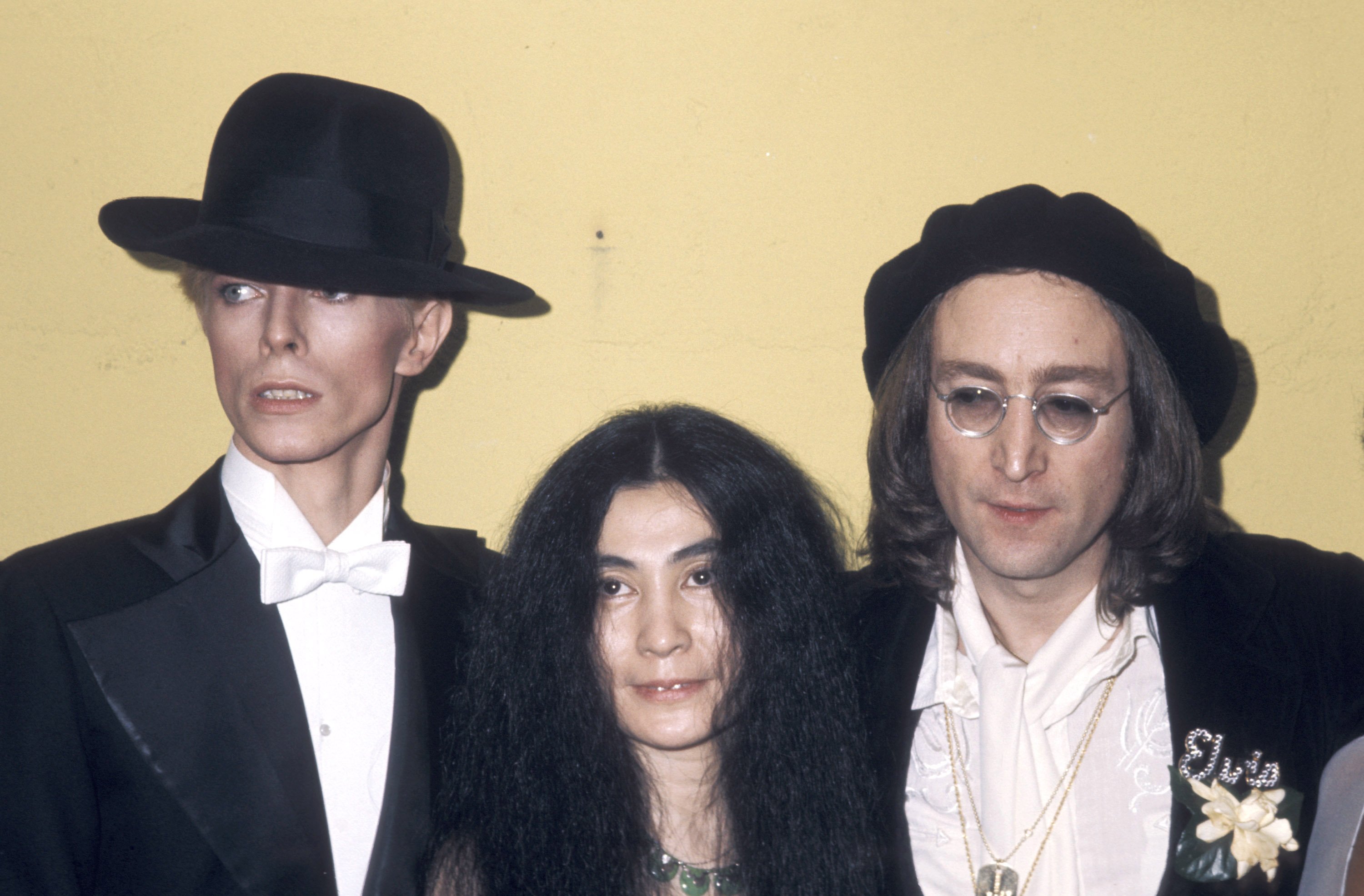 David Bowie, Yoko Ono, and John Lennon wear black and stand in front of a yellow background.