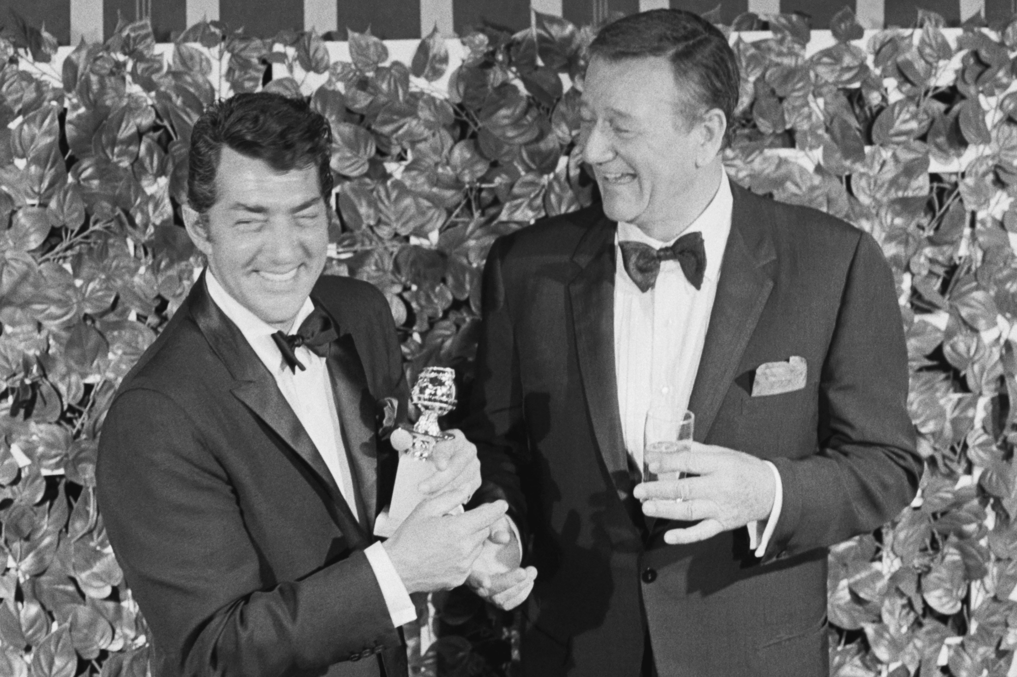 Dean Martin and John Wayne in a black-and-white picture holding a Golden Globe award with both men dressed in tuxes.