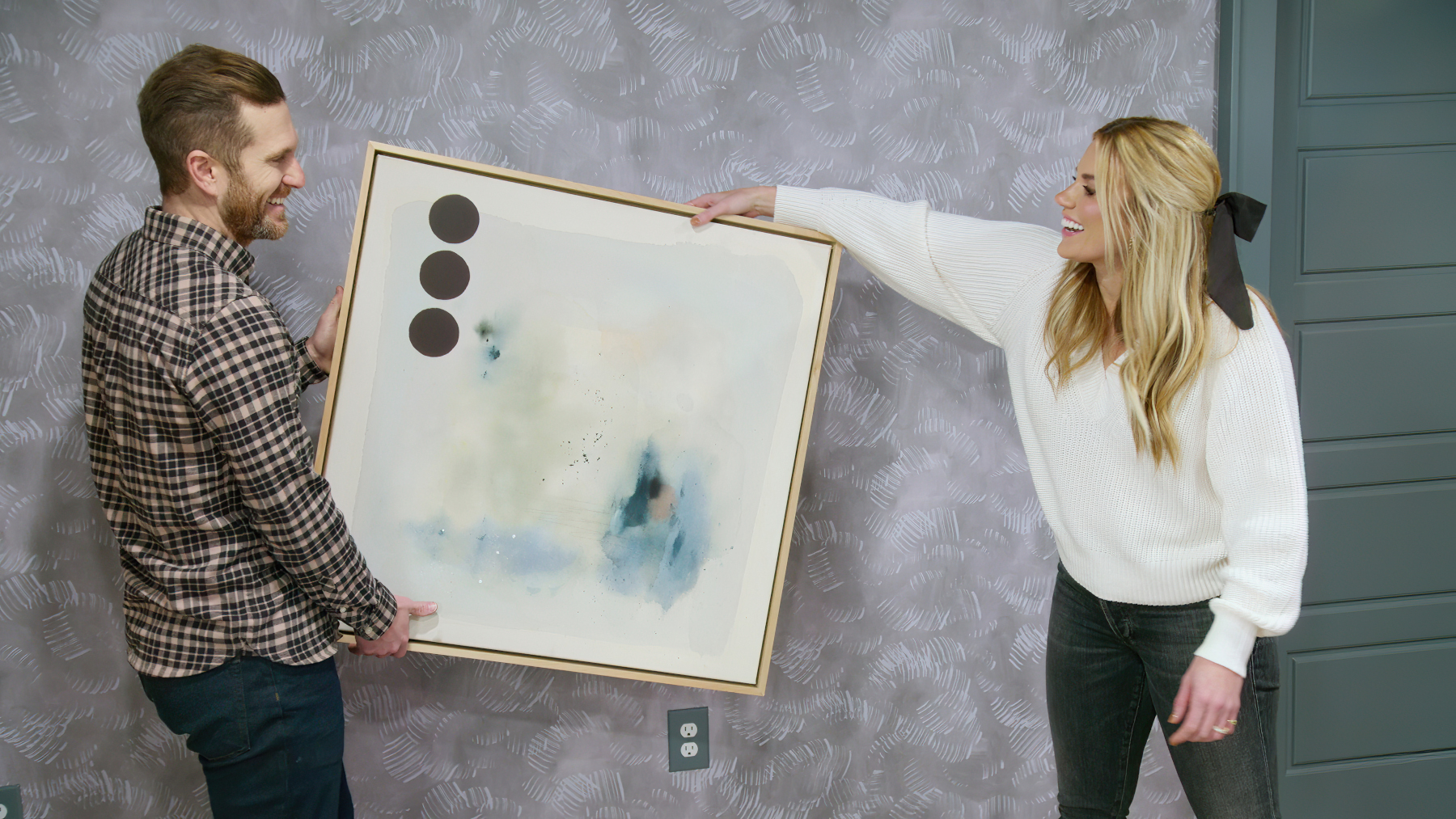Syd and Shea McGee in 'Dream Home Makeover' for our article about the release date of season 4. They're holding a painting, about to put it on a wall.