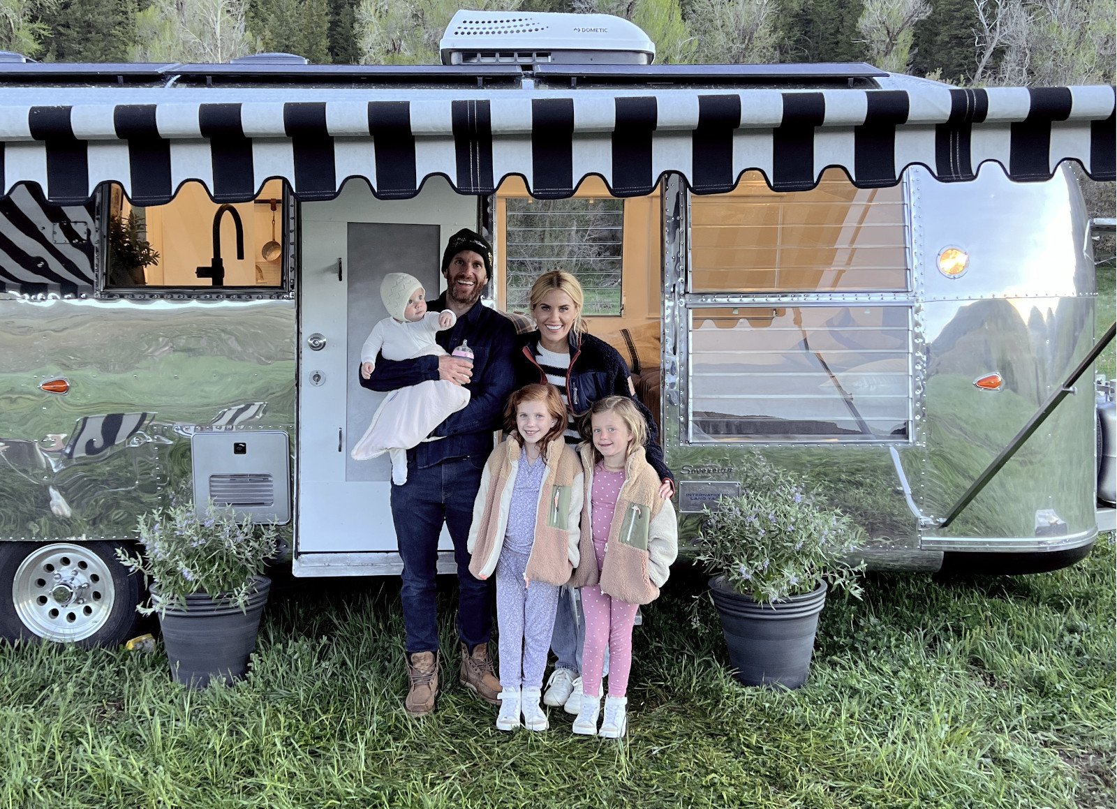 'Dream Home Makeover' Season 4 hosts Shea and Syd McGee and their three daughters. They're standing in front of a trailer and a bunch of trees.