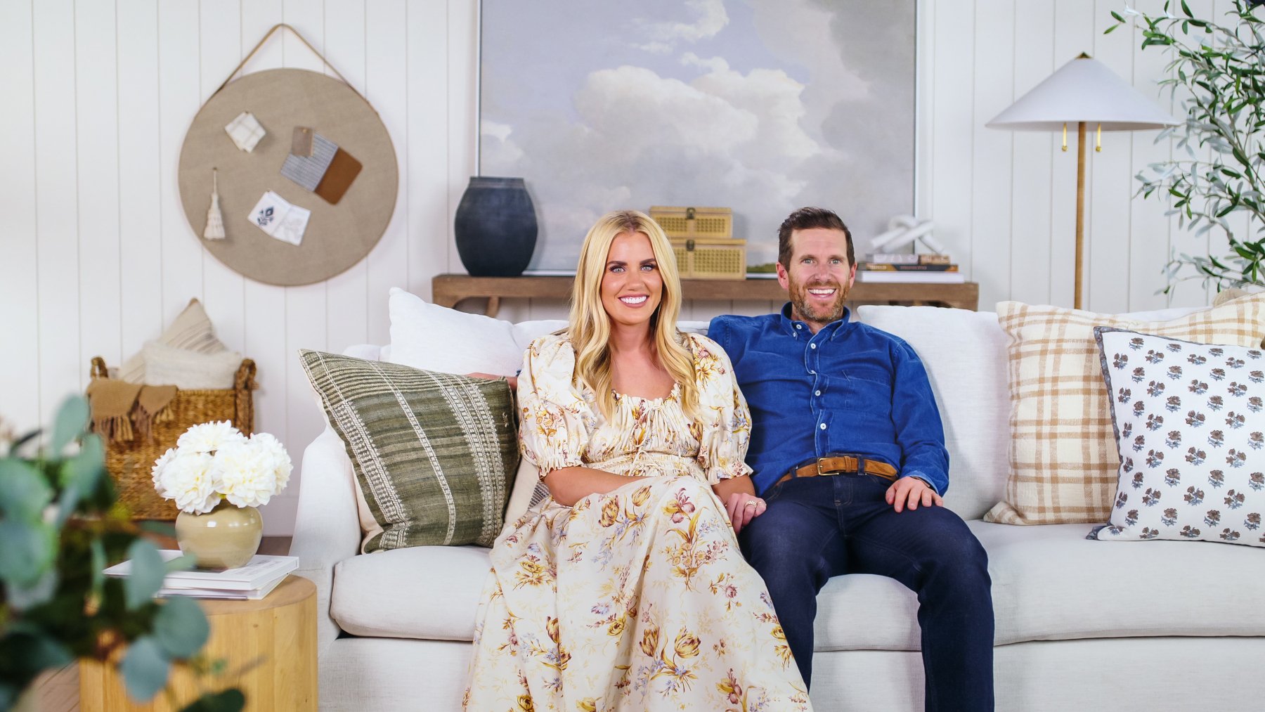 'Dream Home Makeover' hosts Shea and Syd McGee sitting on a couch for our article about their net worth. She's wearing a white, floral dress and he's wearing a blue shirt.