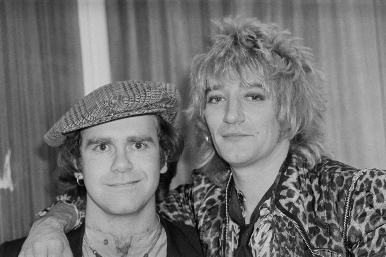 A black and white picture of Rod Stewart with his arm around Elton John's shoulders.
