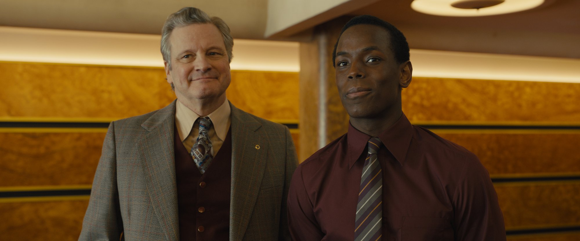 'Empire of Light' Colin Firth as Donald Ellis and Micheal Ward as Stephen. They're standing next to each other wearing suit and ties smiling in front of wall with wooden panels behind them