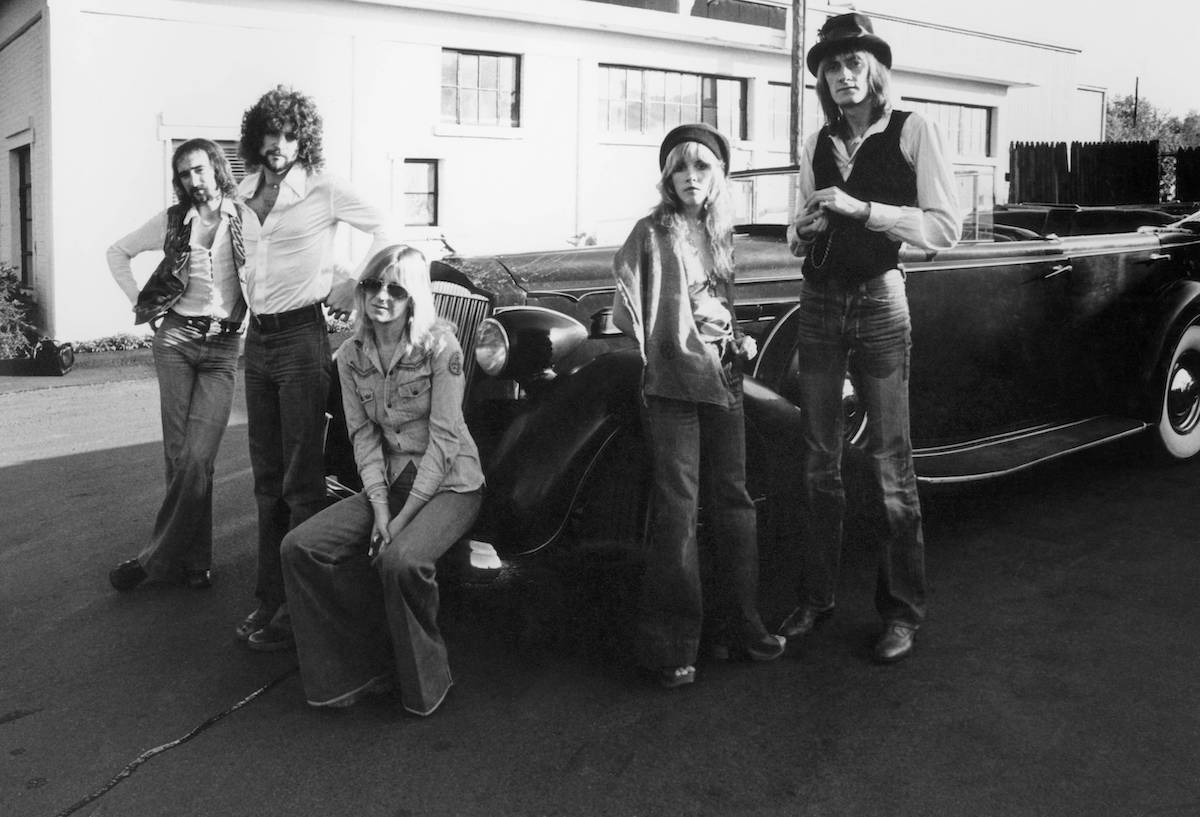 A black and white photo of Fleetwood Mac members John McVie, Lindsey Buckingham, Christine McVie, Stevie Nicks, and Mick Fleetwood posing around a car.