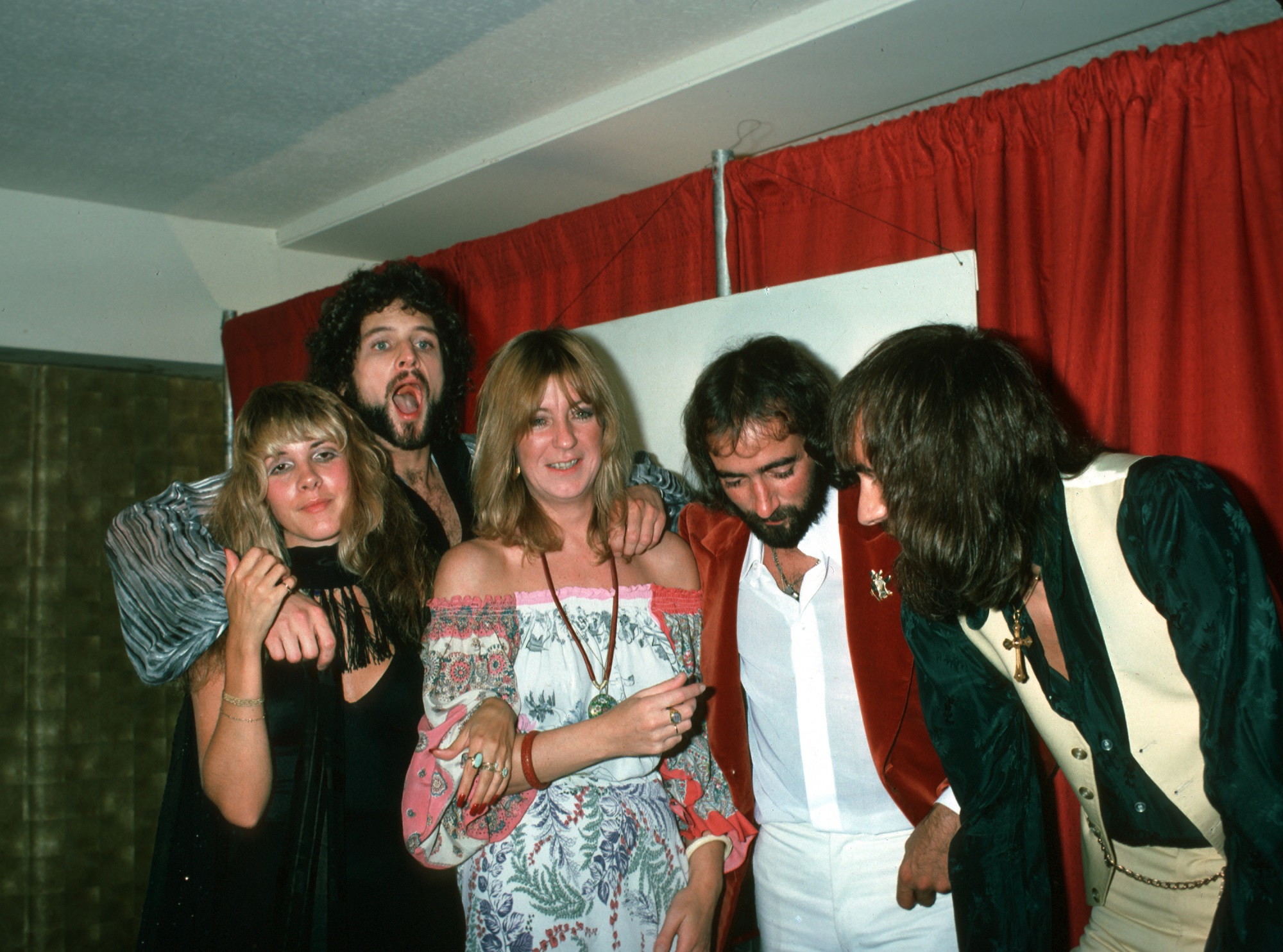 Stevie Nicks, Lindsey Buckingham, Christine McVie, John McVie and Mick Fleetwood of Fleetwood Mac huddle together in front of a red curtain backstage